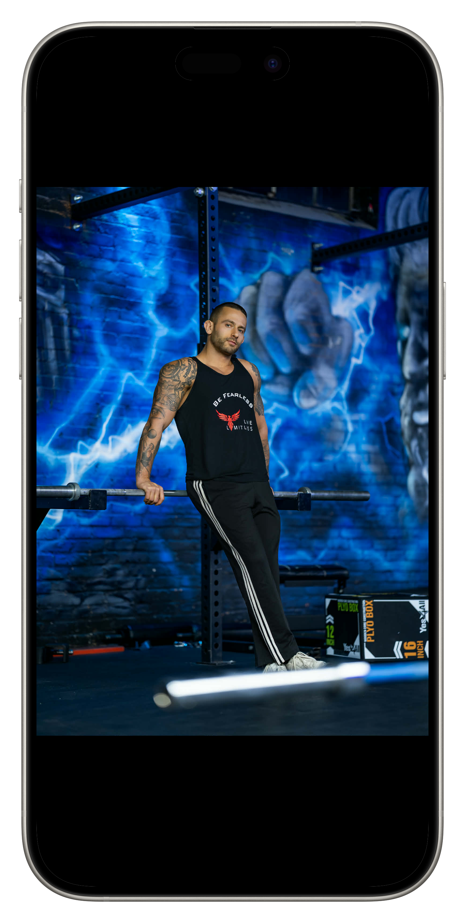A man with tattoos on his arms wearing a black tank top and black track pants with white stripes, standing in a gym leaning on a barbell with a blue brick wall and graffiti artwork in the background.