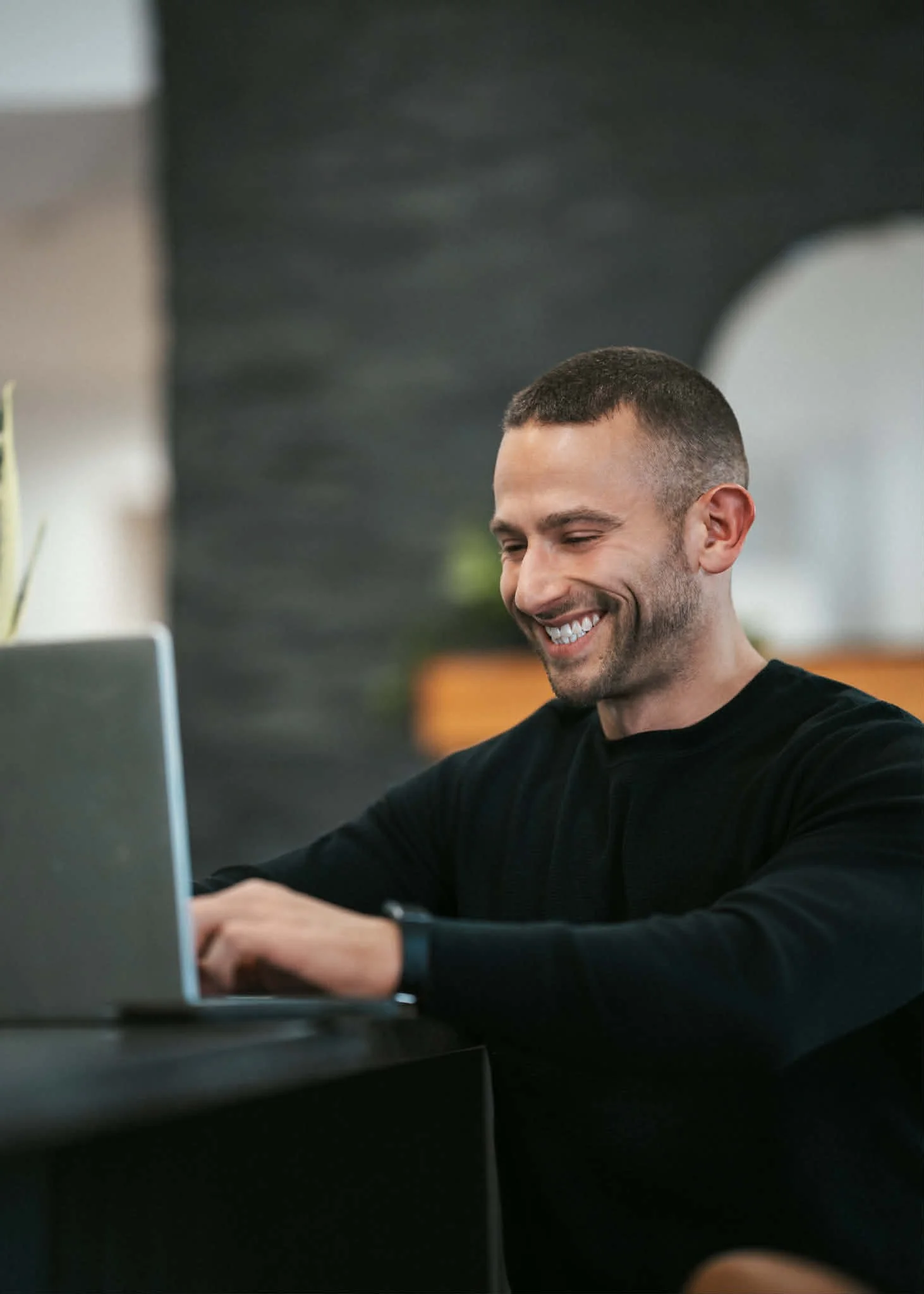 A man with a short haircut and a beard, wearing a black long-sleeve shirt, sitting at a desk and smiling while working on a laptop.