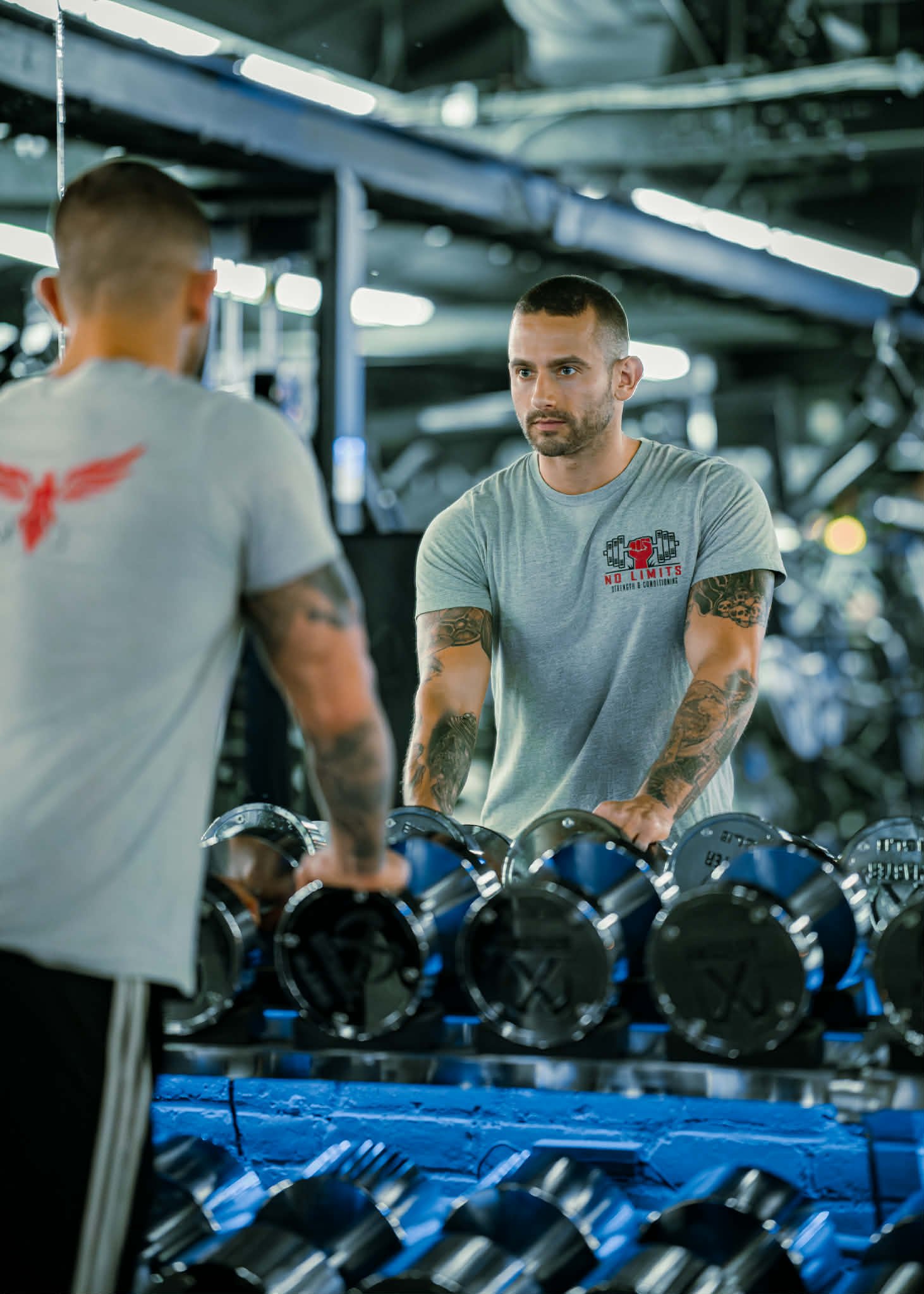 Two men in a gym looking at a row of dumbbells on a rack, with one man facing the camera and the other with his back to the camera. The man facing the camera has tattoos on his arms, short hair, and is wearing a gray T-shirt with a red and black logo.