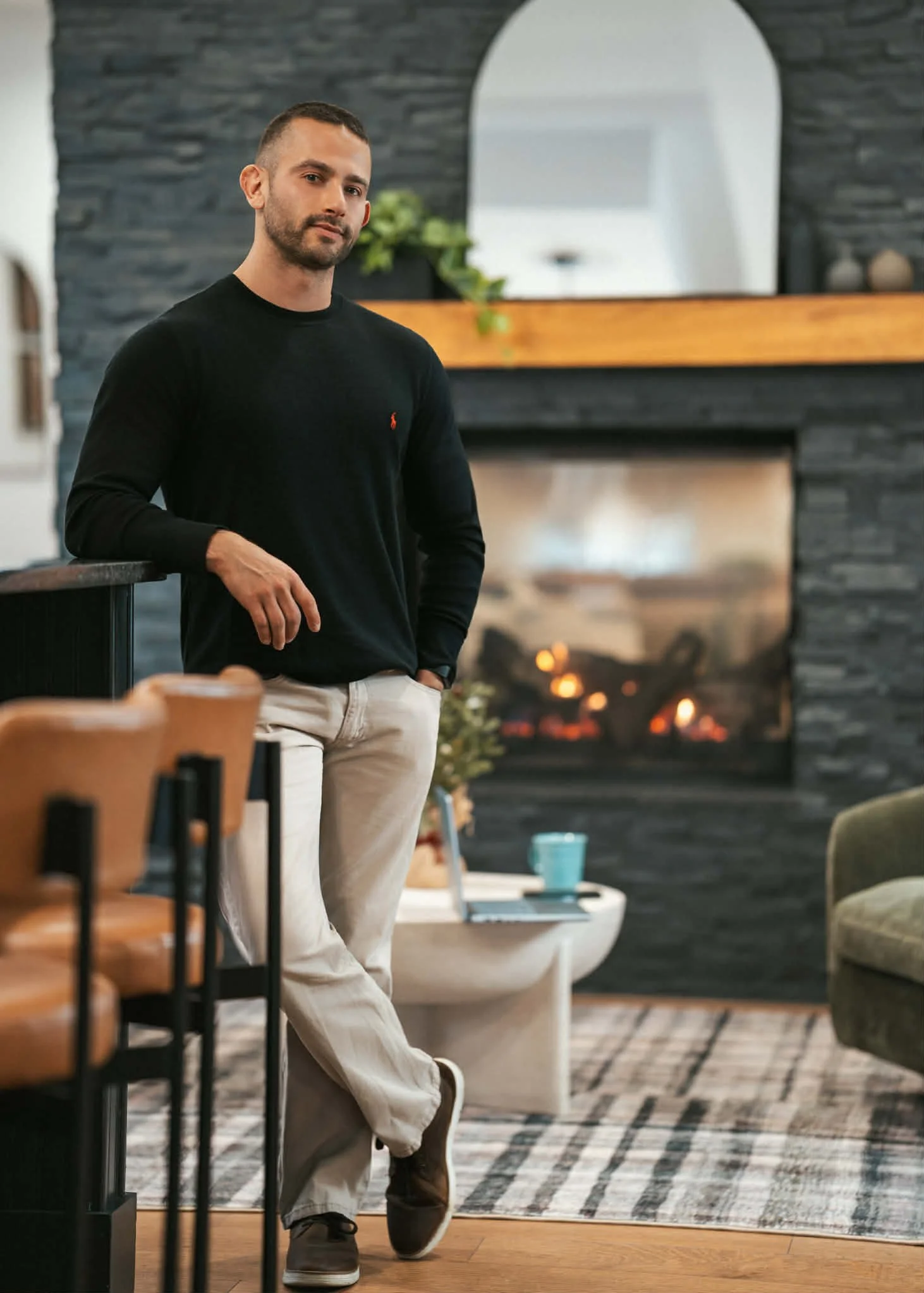 A man with short dark hair and a beard stands in a living room with a fireplace, wearing a black long-sleeve shirt and beige pants.