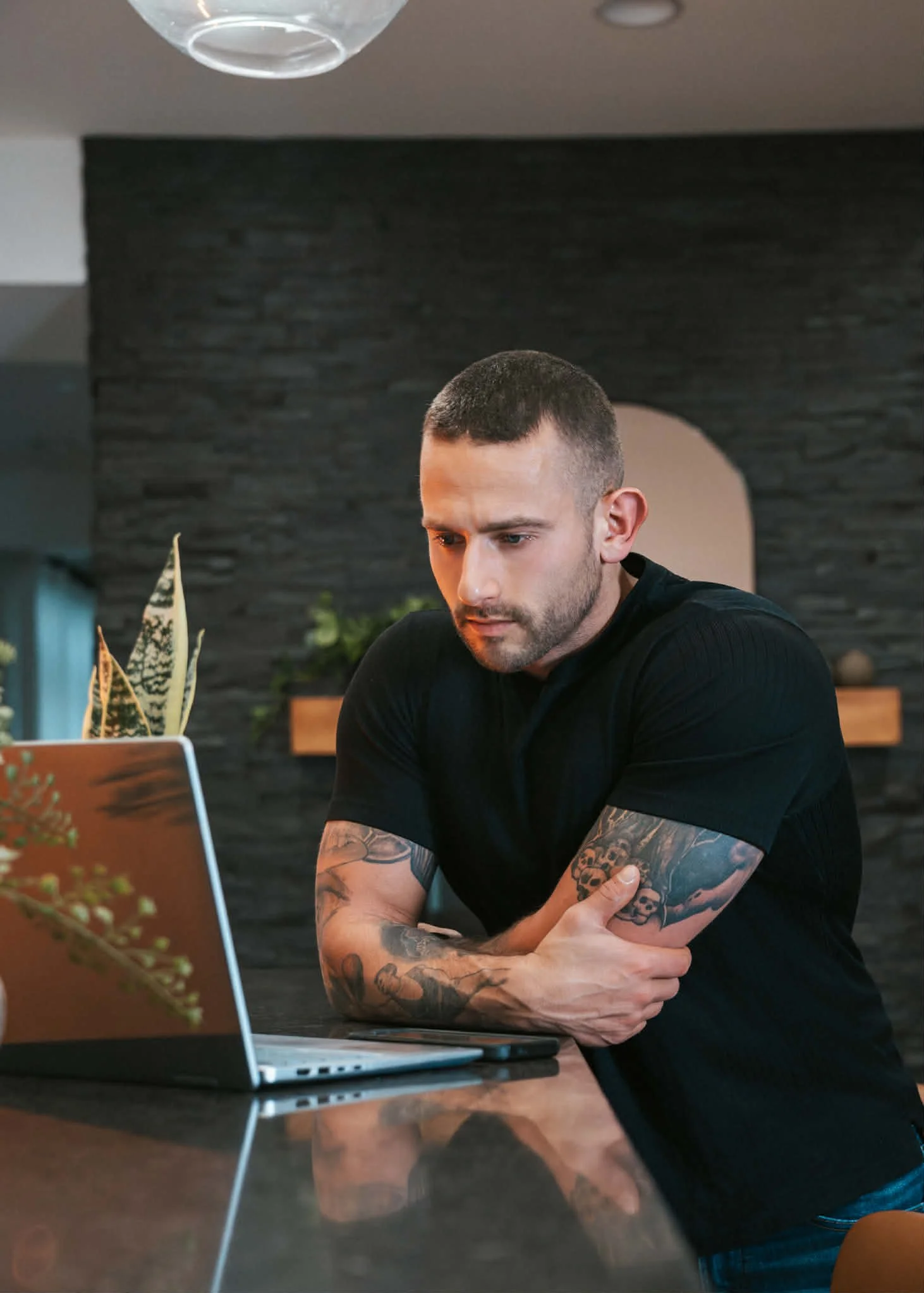 A man with tattoos on his arms and short hair sits at a table with a laptop, appearing focused on his work.
