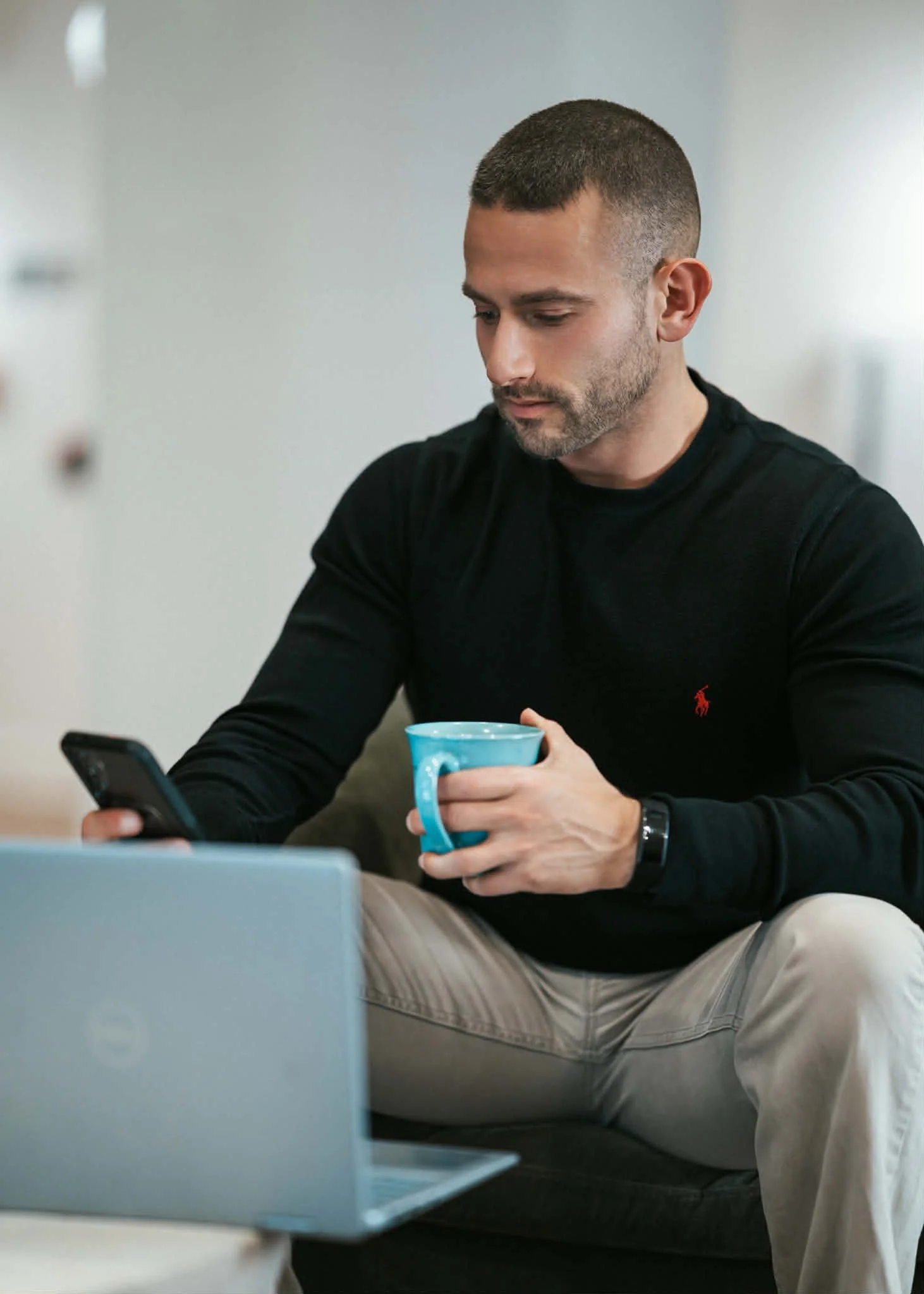 A man with short hair and a beard, wearing a black shirt, is sitting on a sofa, looking at his phone, holding a blue mug. There is a laptop and a wall in the background.