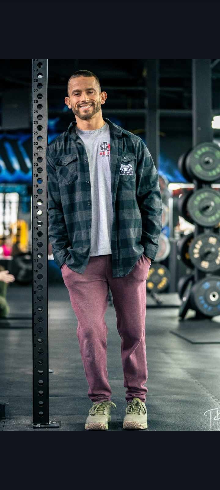 A smiling man standing in a gym next to a measuring pole, wearing a plaid shirt, gray T-shirt, pink sweatpants, and sneakers.