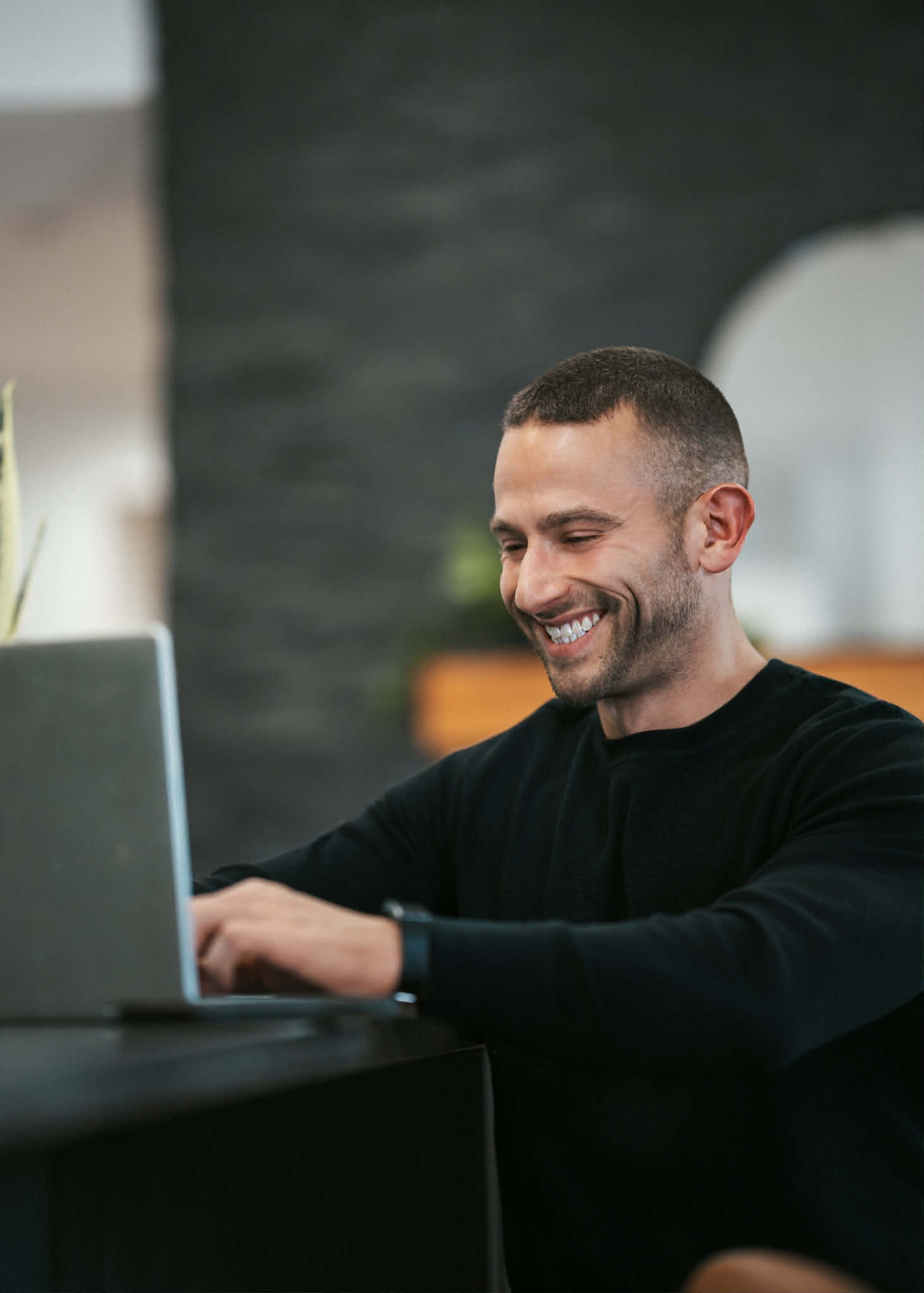 A man with short hair and a beard smiling, working on a laptop at a desk.