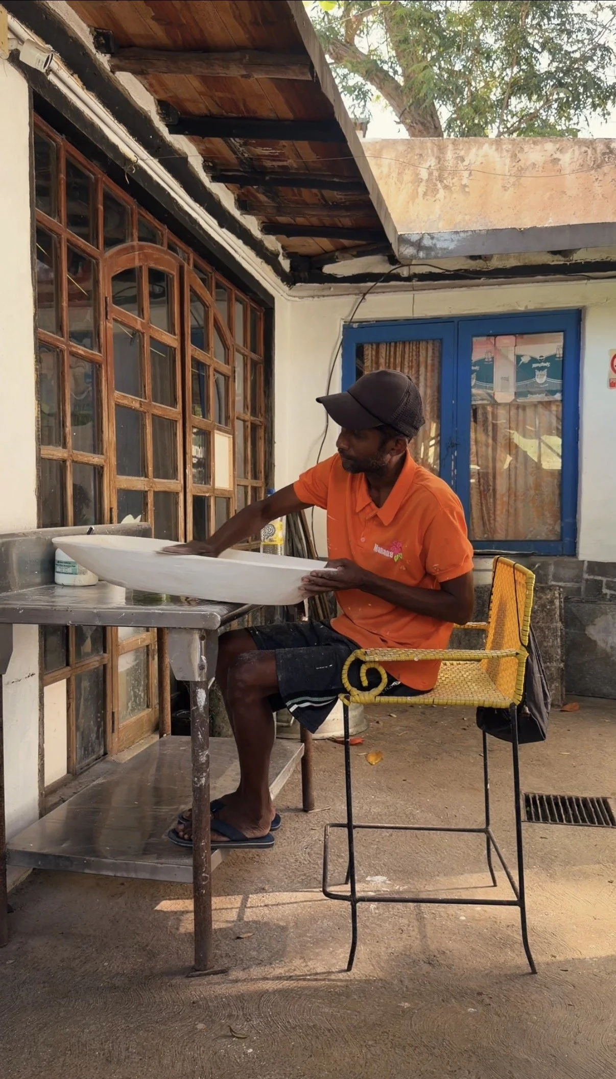 A man wearing an orange shirt, black shorts, and a cap is sitting on a yellow woven chair outdoors. He is cleaning a white surfboard on a metal table. Behind him, there is a large window with wooden panes, a blue door with a window, and a building wi