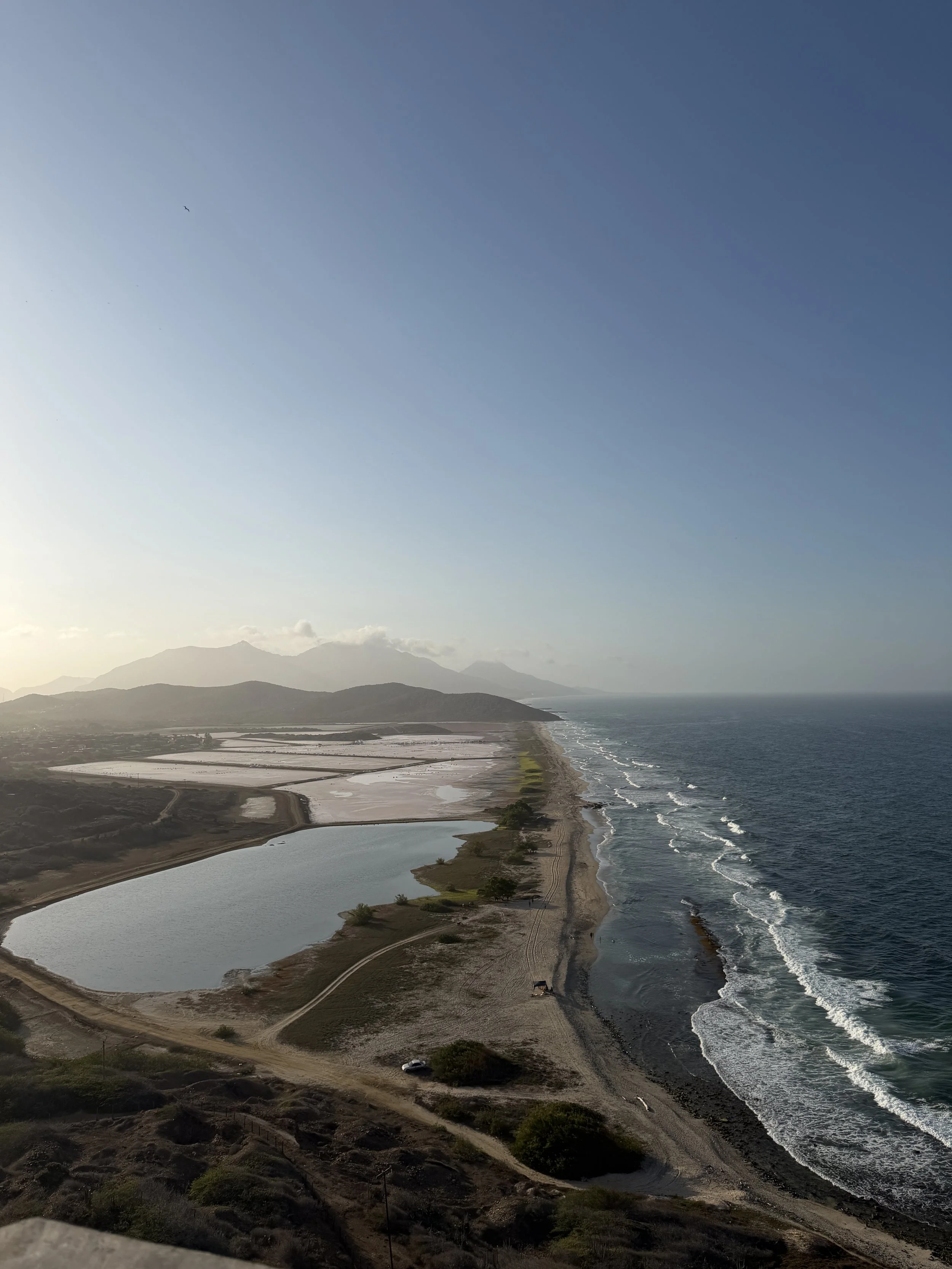 A coastal landscape with a beach, a body of water, and mountains in the background. The sky is mostly clear with a small bird visible in the upper left.