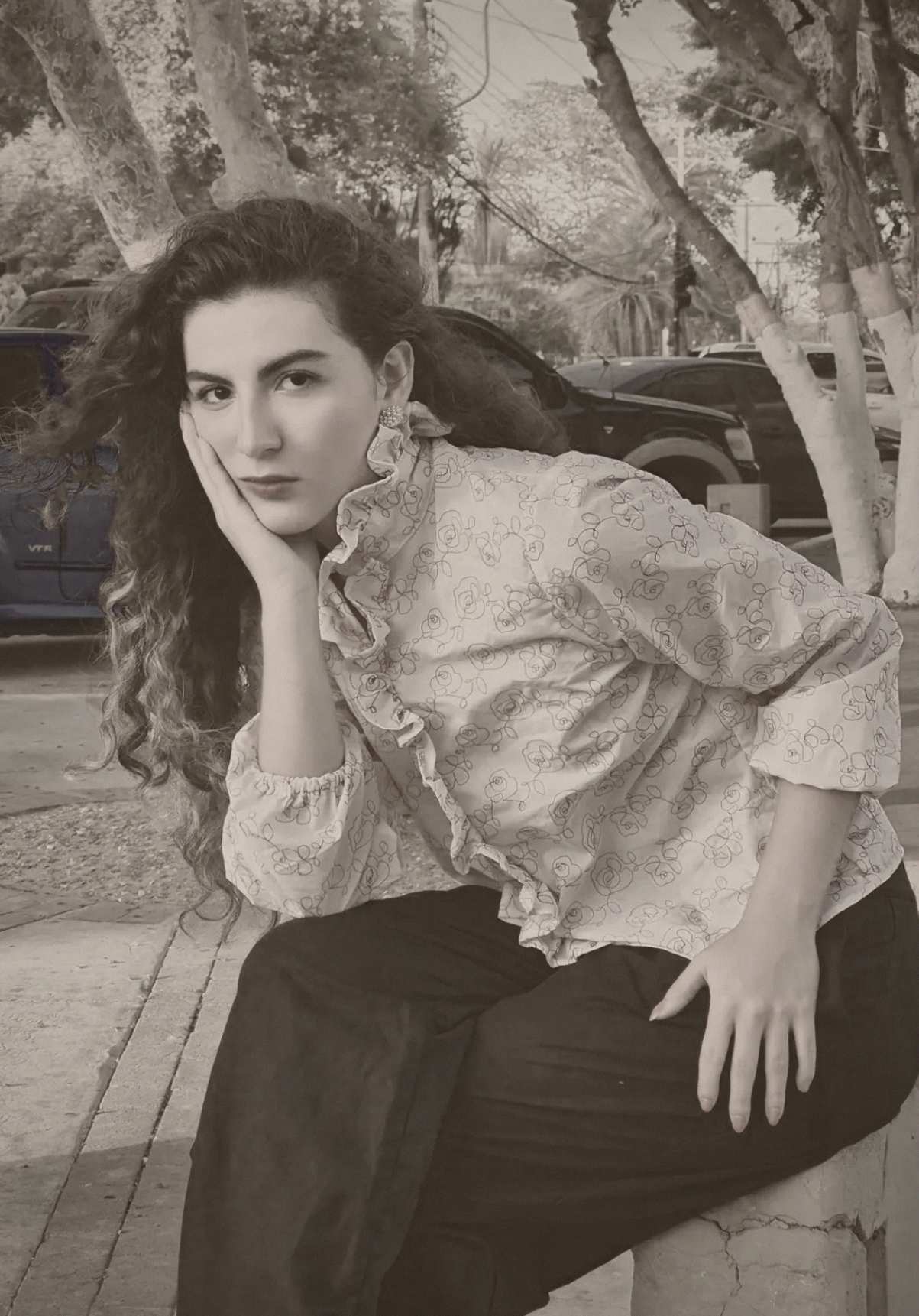 A black and white photo of a woman with curly hair, wearing a patterned blouse with ruffles, sitting outdoors on a curb with trees and parked cars in the background.