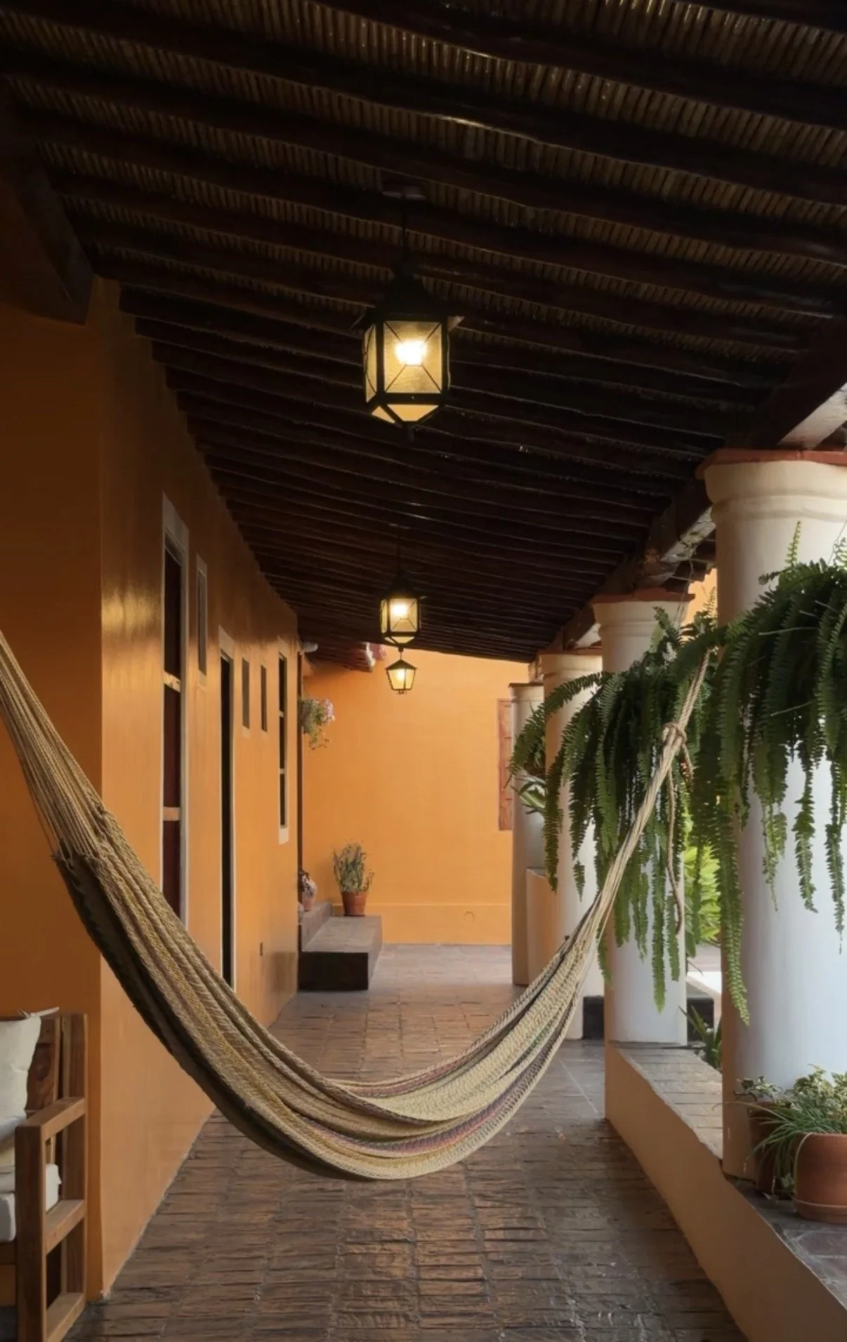 Covered porch with yellow walls, hanging lantern lights, potted plants, a hammock, and white columns supporting a dark wooden ceiling.