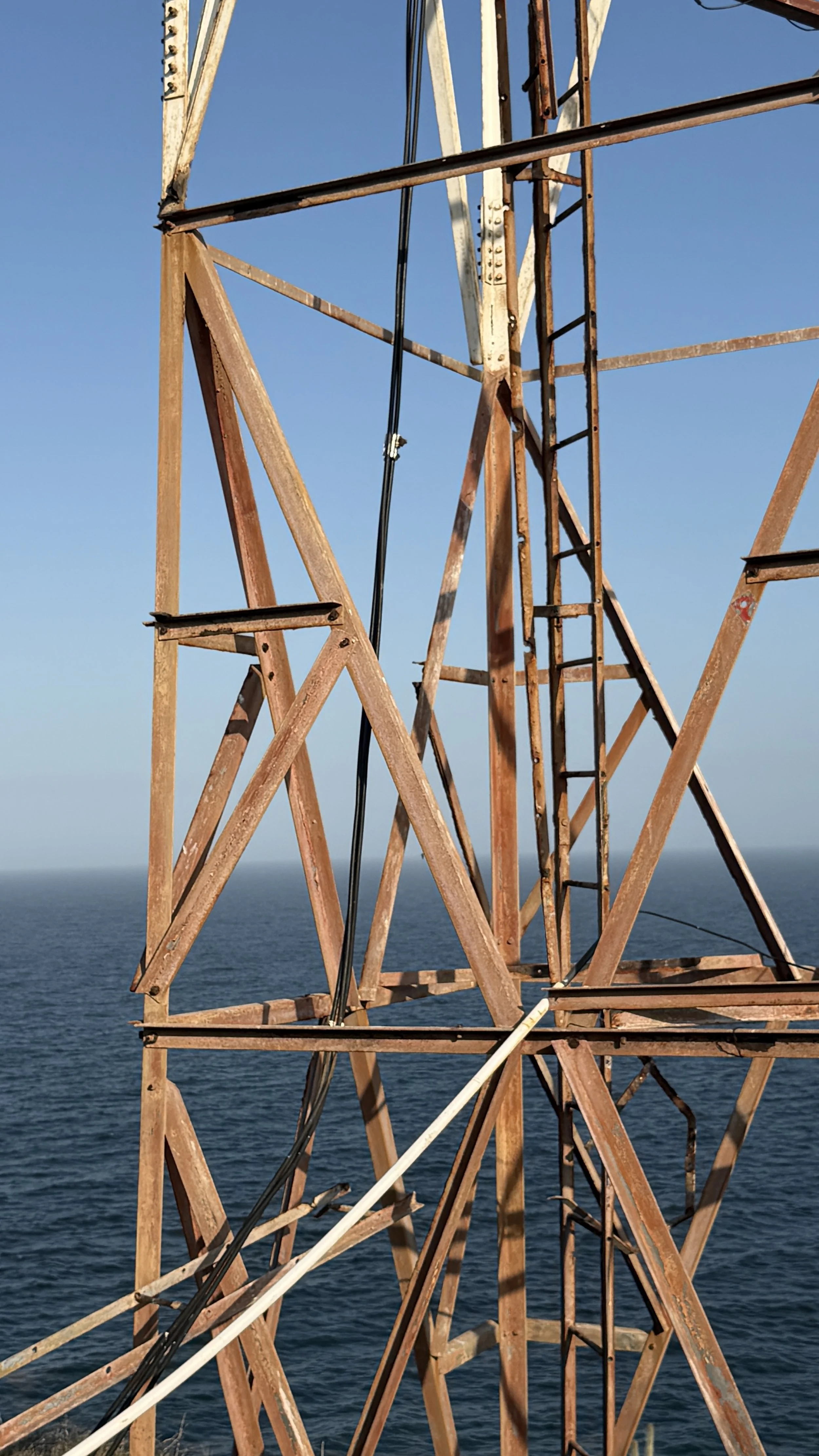 Close-up of a rusty metal structure of a tower with the ocean in the background and a clear blue sky.
