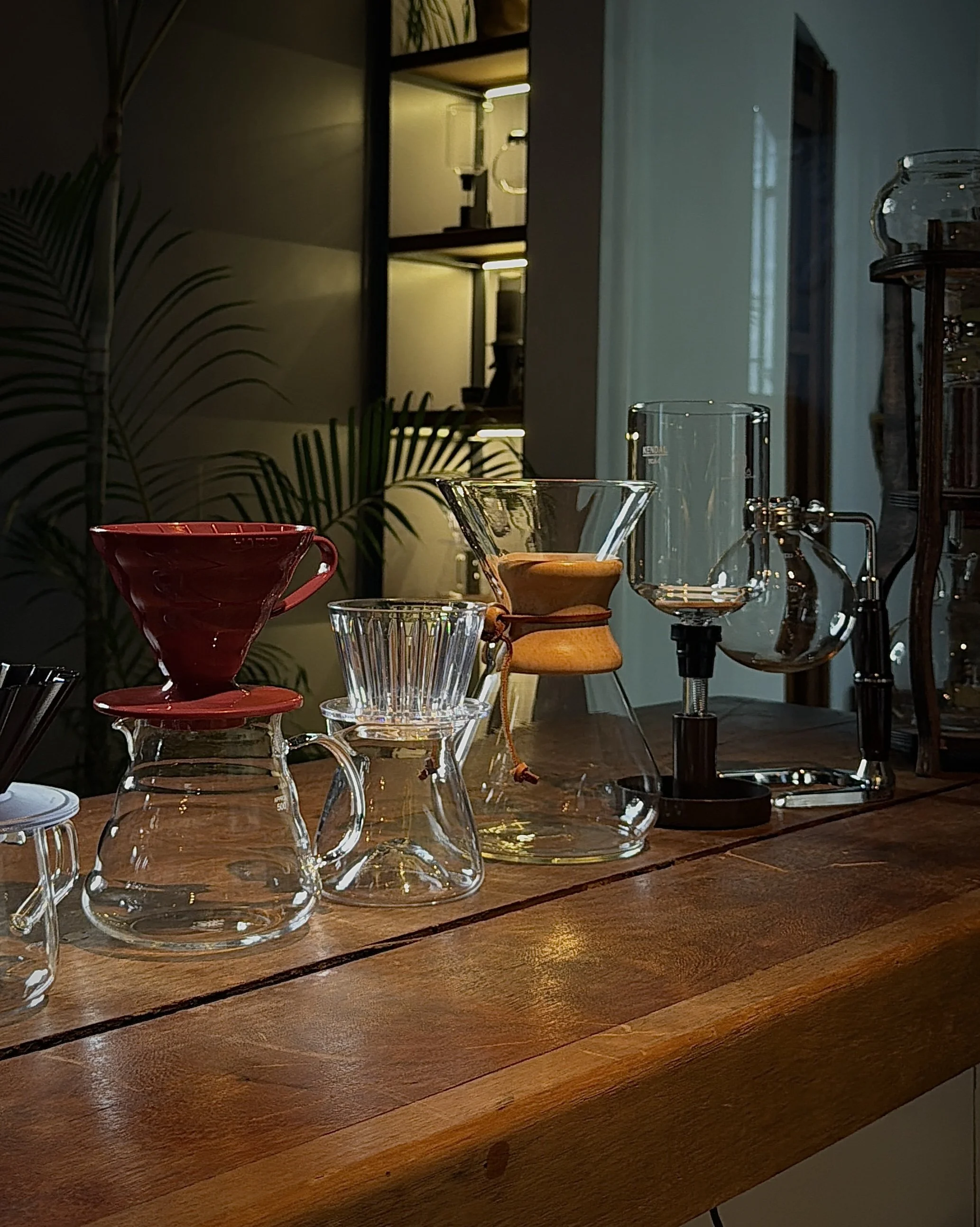 Close-up of a wooden table displaying various glassware, including a red bowl, a clear glass with a textured pattern, a funnel-shaped glass, and other laboratory glassware. In the background, a shelf with custom lighting, artwork on the wall, and a g