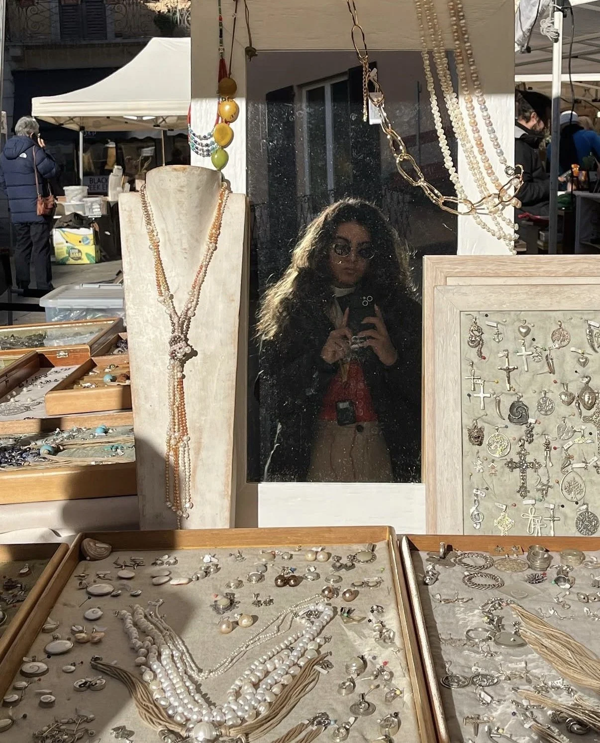 Outdoor jewelry market stall displays necklaces, earrings, and charms with a mirror reflecting a woman taking a photo, with tents and people in the background.