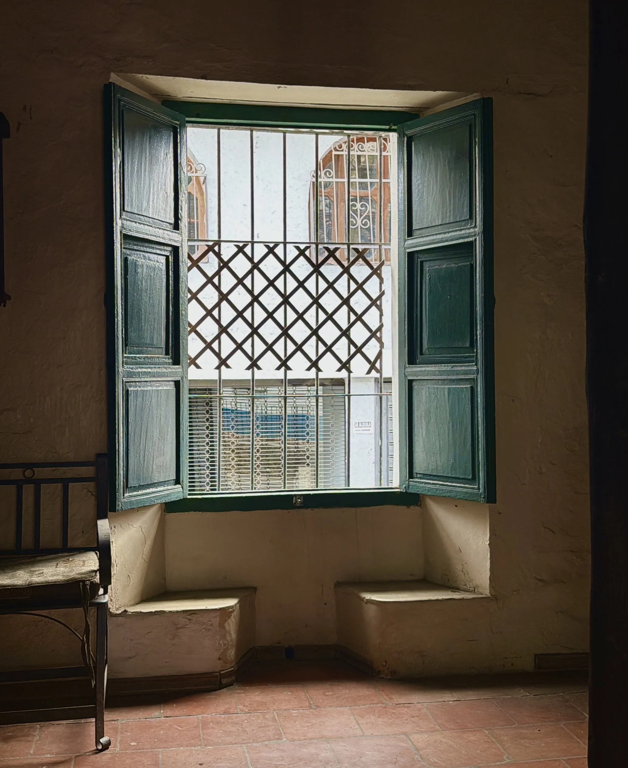 A view from inside a building looking out a window with green shutters open, showing a small balcony with metal bars and a lattice gate, with a street scene and another building with ornate window frames in the background.