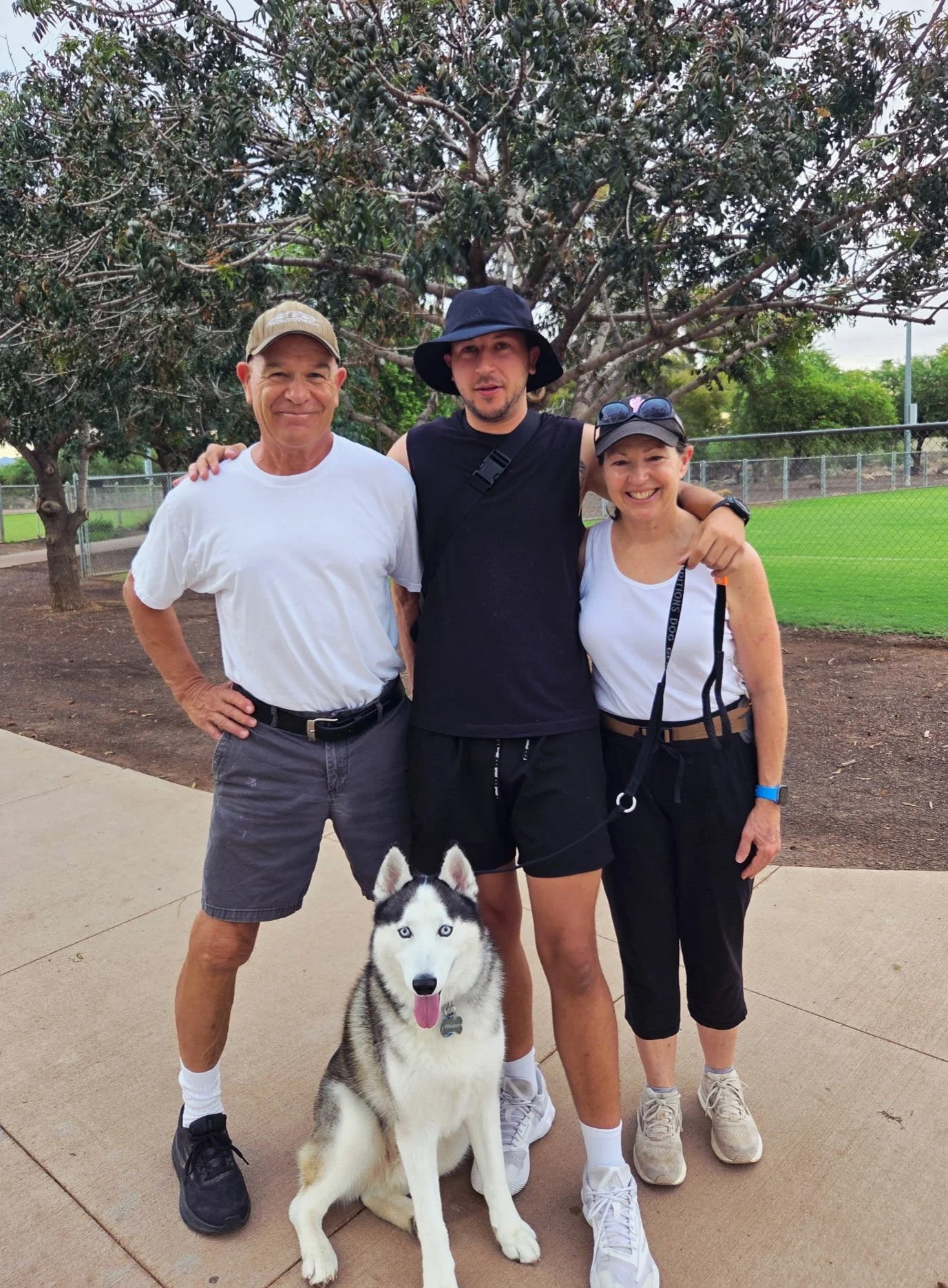 Three people and a husky dog standing outdoors on a sidewalk with a grassy field and trees in the background. The group includes one man wearing a white t-shirt and a cap, a man in a black sleeveless top and hat, and a woman in a white tank top and cap. The husky dog is sitting in front of them, facing the camera.