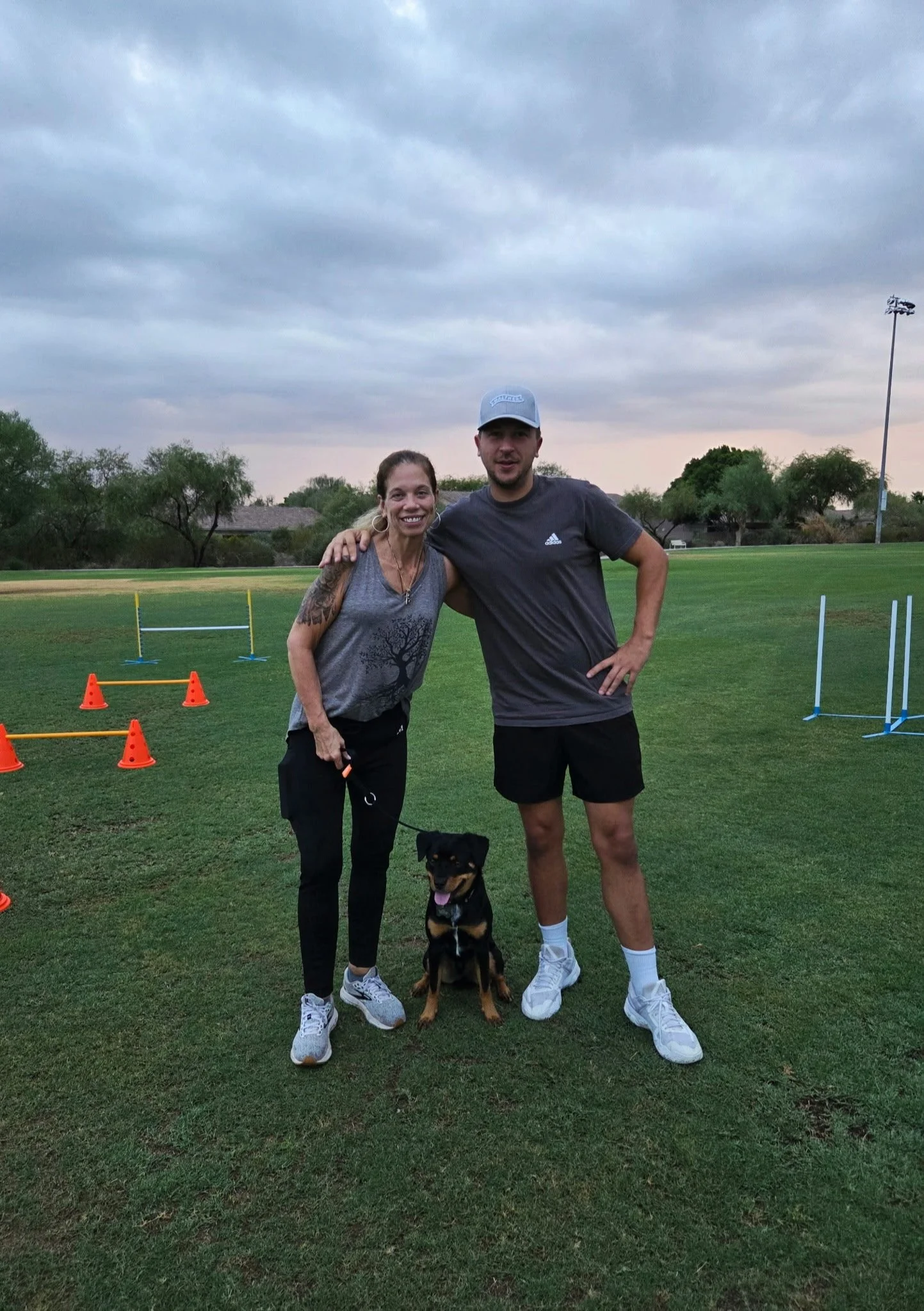 A woman, a man, and a dog standing together on a grassy field, with agility training poles and cones set up behind them, under a cloudy sky.