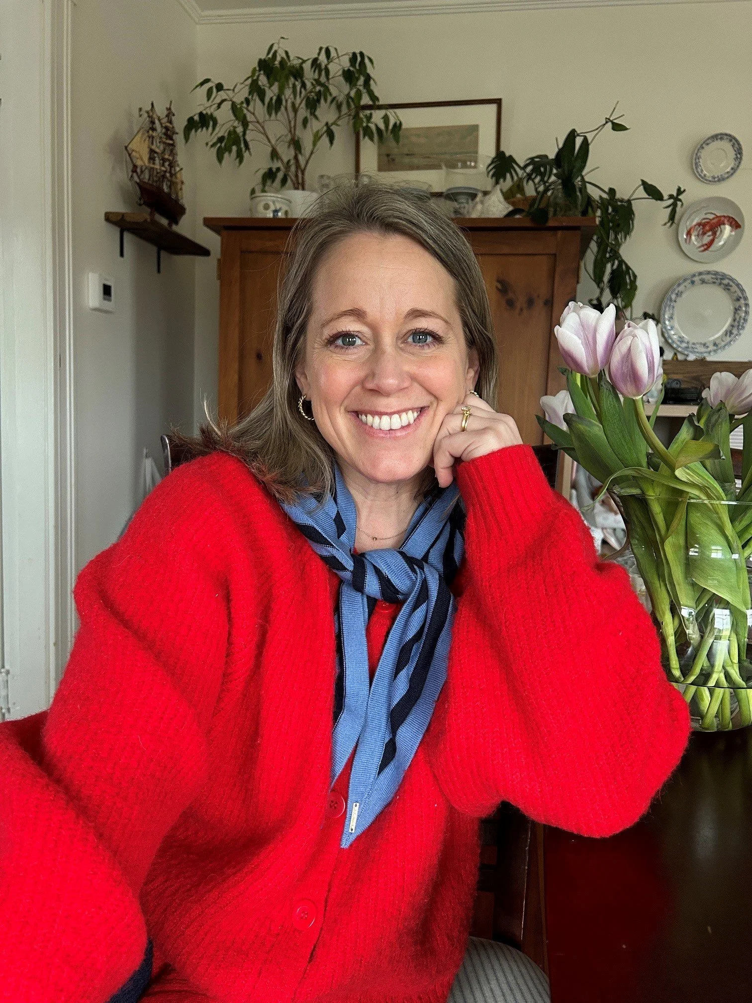 A smiling woman with blonde hair wearing a red cardigan and blue and black striped scarf, sitting at a table with a vase of pink tulips in front of her.