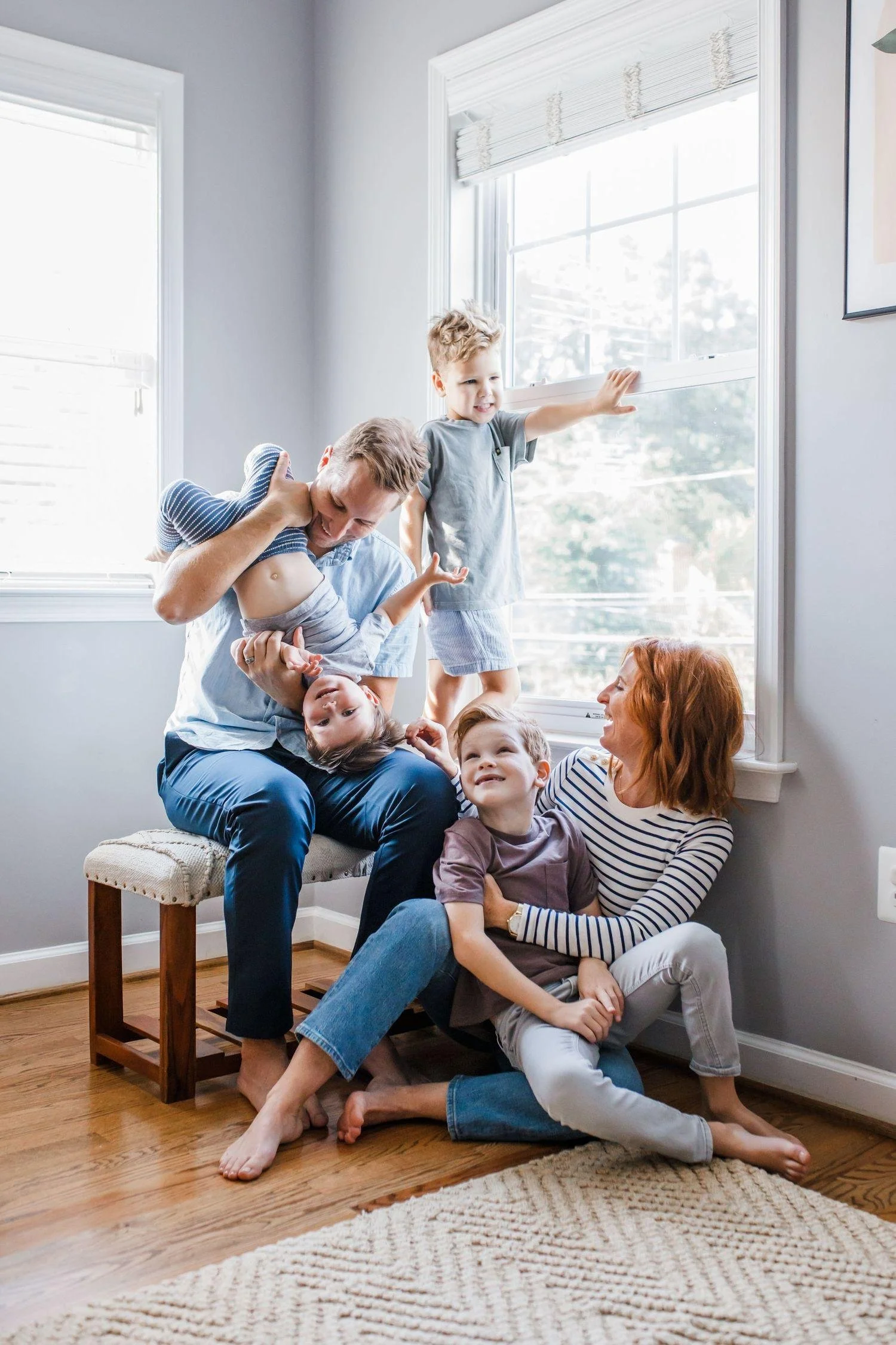 A family of six enjoying a playful moment together in a bright living room with large windows, hardwood floors, and a beige rug. The group includes children and adults, with one adult holding a child upside down and another adult sitting on the floor with children on her lap.