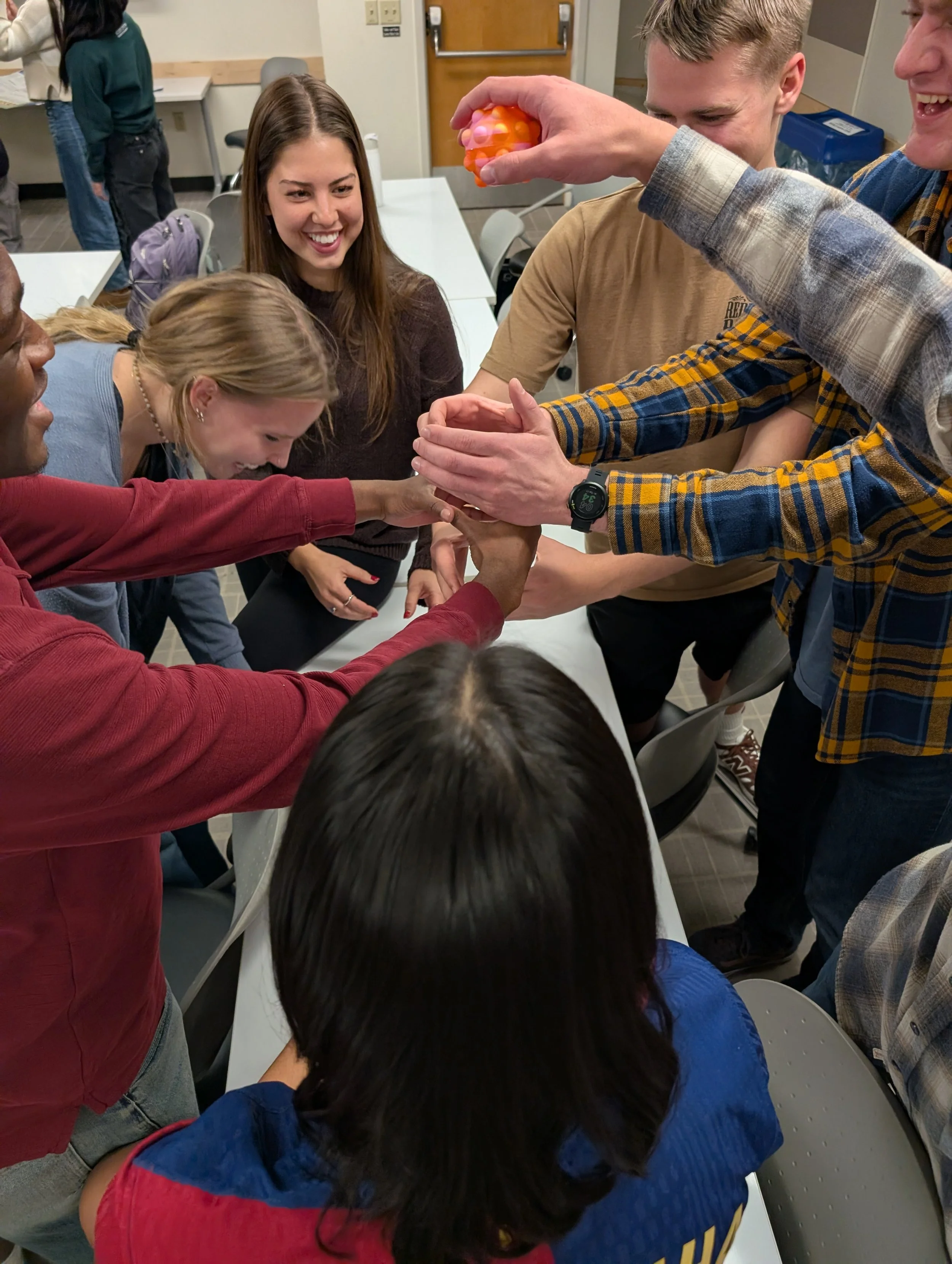 A group of people gathered around a table, with one person holding a textured orange ball with pink lights, and others reaching and smiling, appearing to be engaged in a game or activity in a room with office doors and tables.