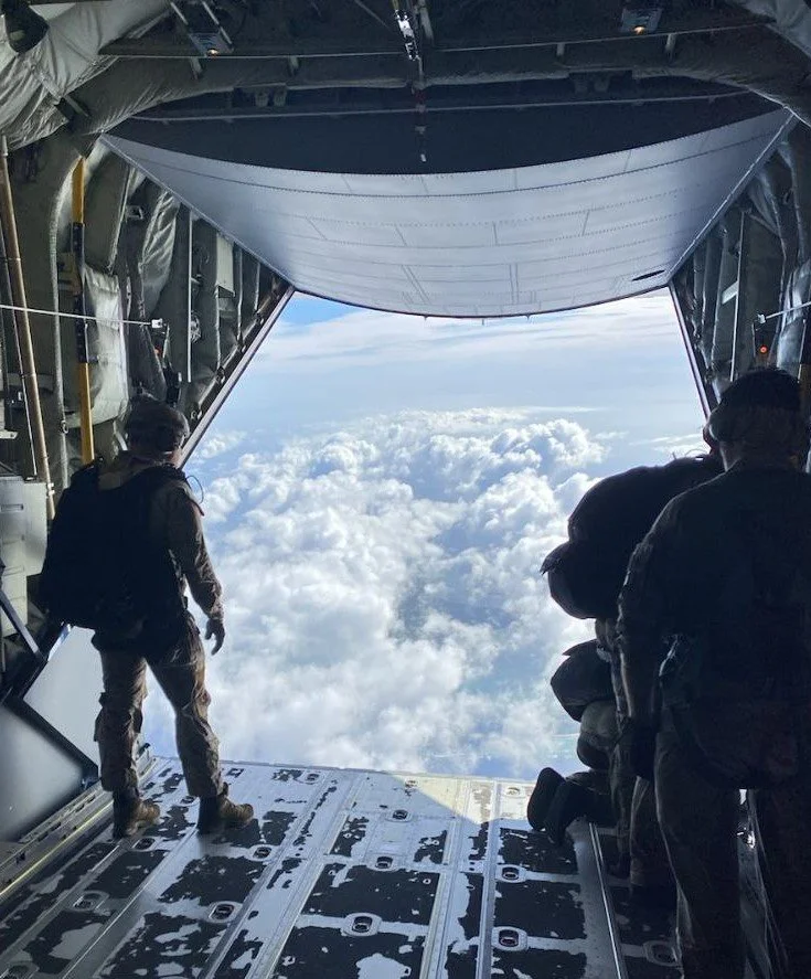 People look out from the open aft cargo door of a military aircraft flying above the clouds.