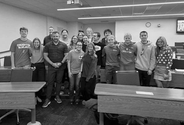 Group of diverse young adults posing together in a classroom or conference room with desks and chairs, some smiling and wearing casual clothing.