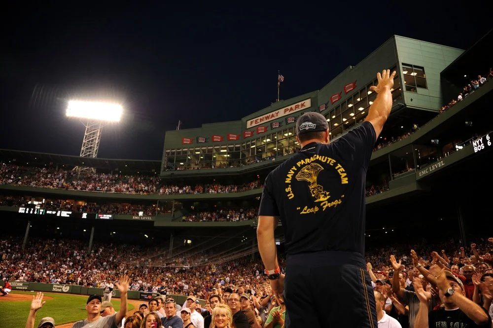 A person in U.S. Navy parachute team attire waving to a crowd at Fenway Park baseball stadium during a night event.