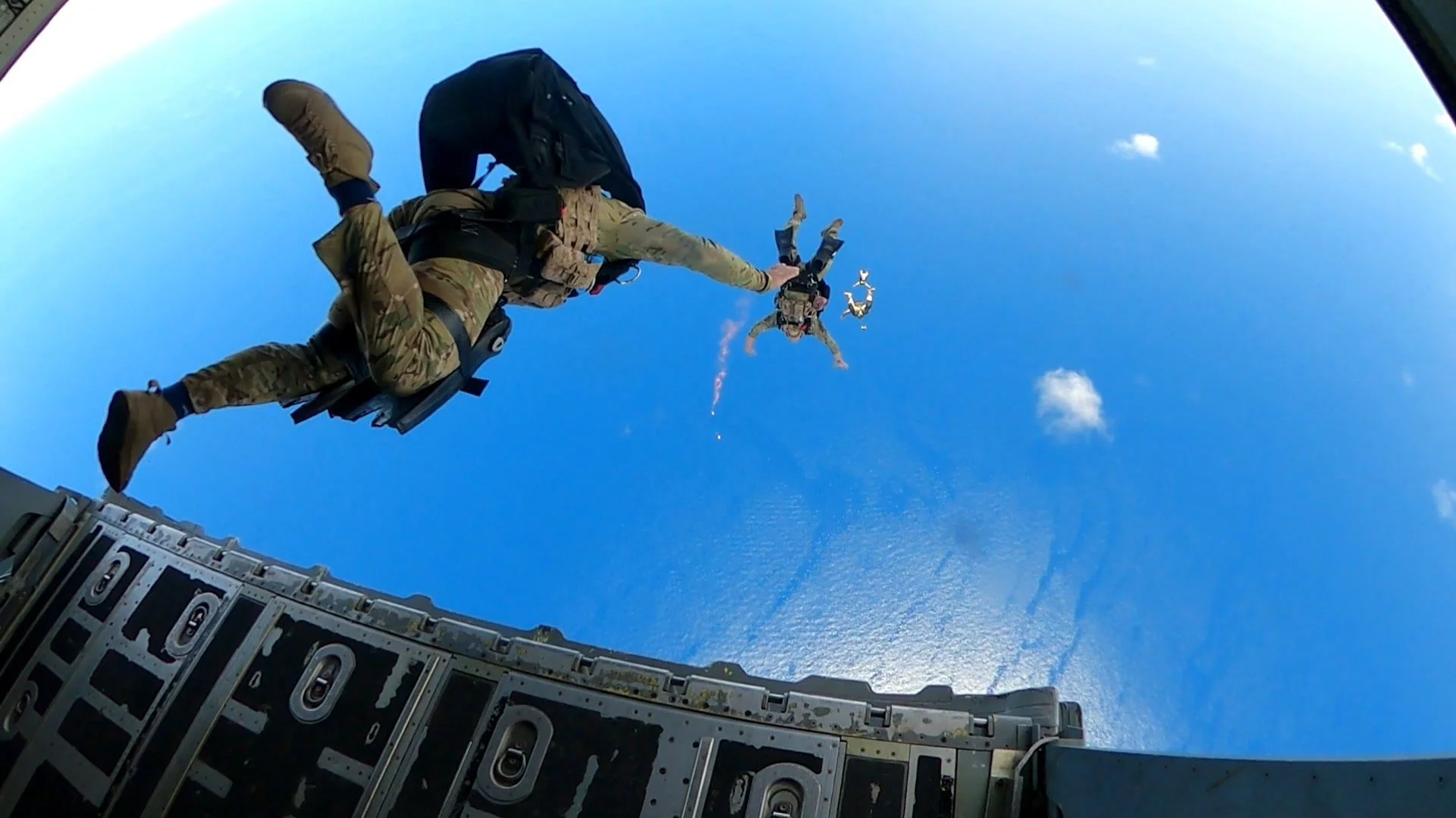 Two soldiers in military gear and parachutes jumping out of a cargo plane with the blue sky and a few clouds in the background.
