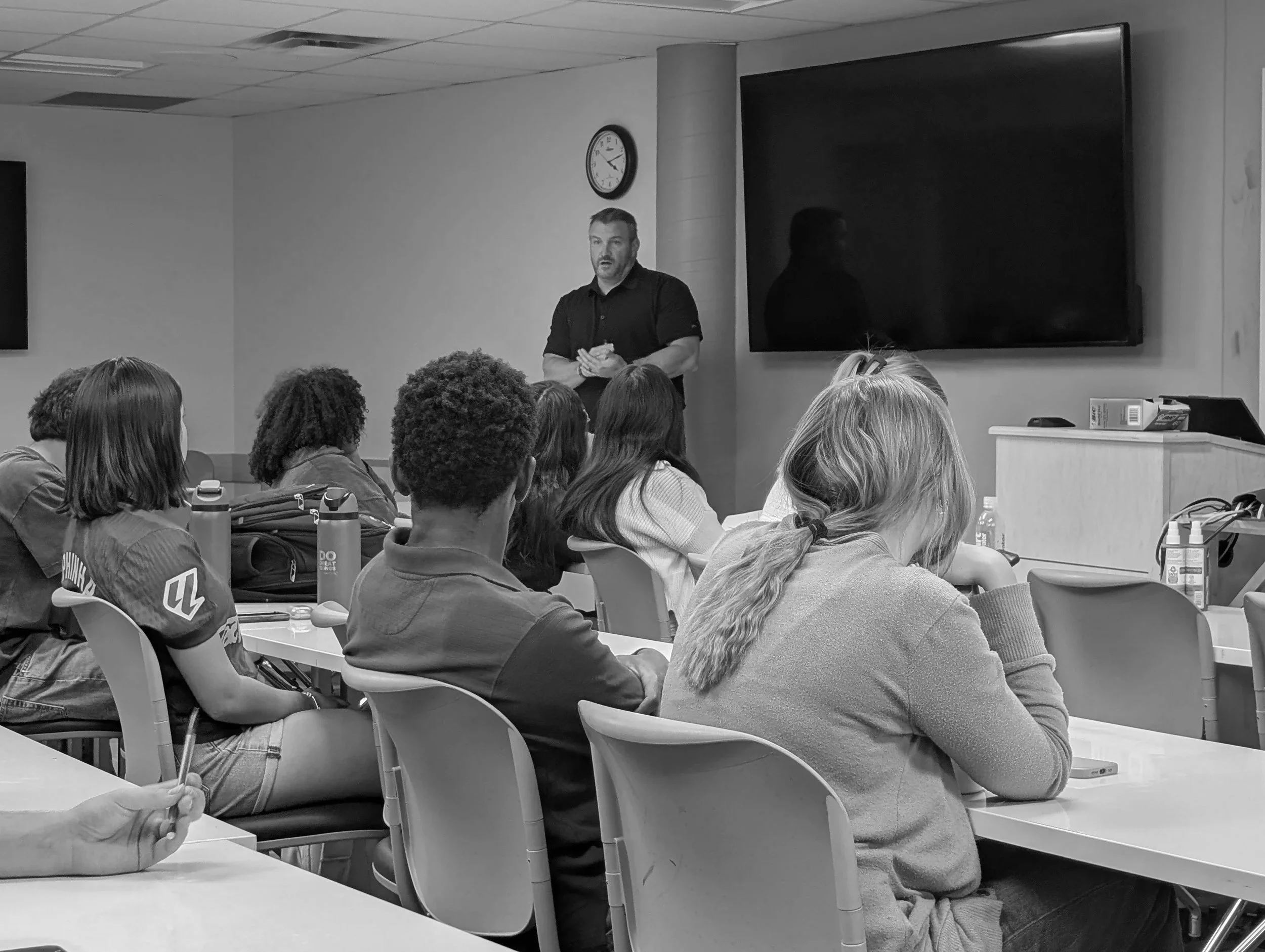 A man in a black shirt standing in front of a group of people in a classroom or meeting room, with a large flat screen monitor and a clock on the wall.