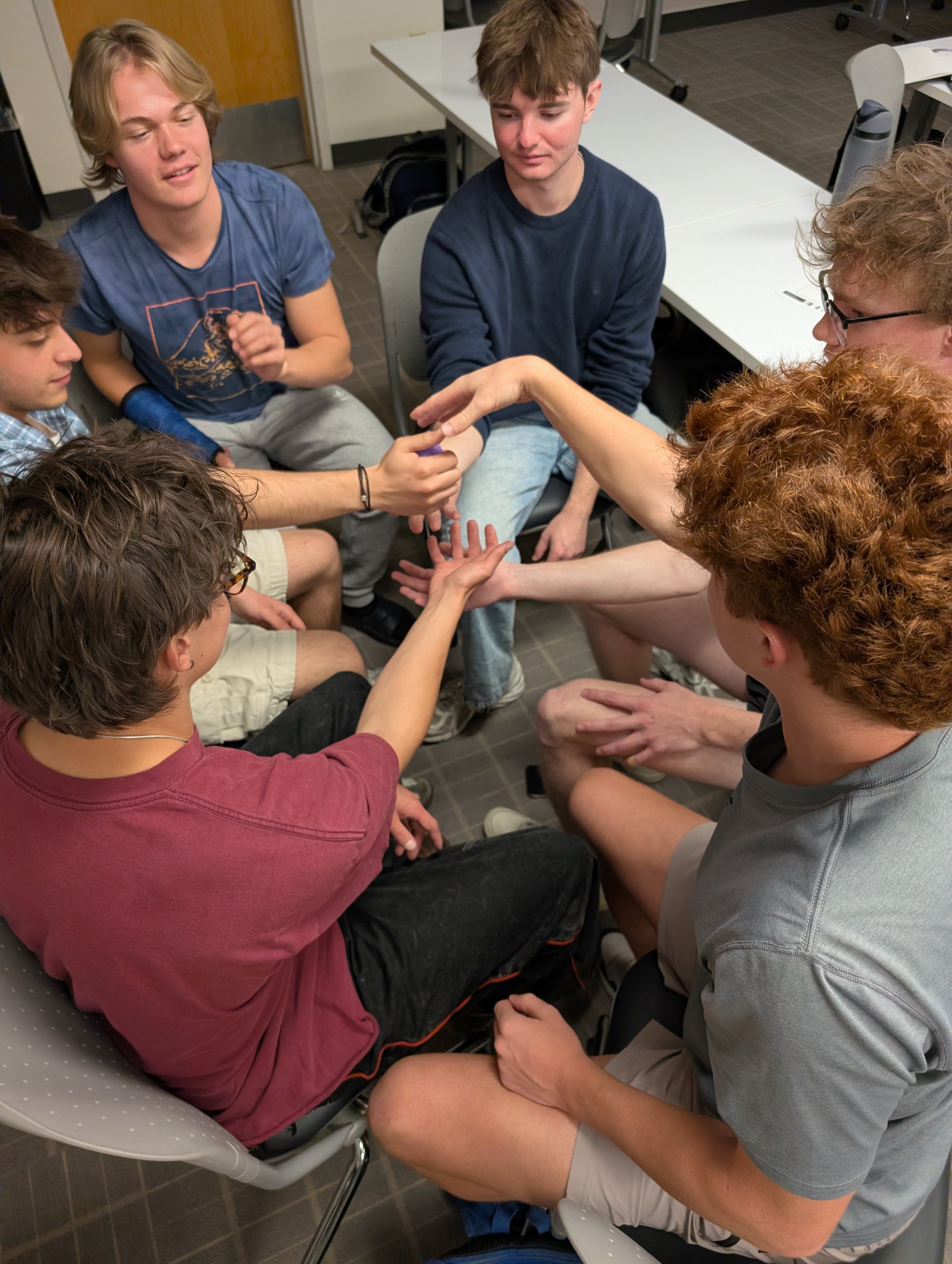 Group of young people sitting closely together in a circle, involved in a handshake activity in a classroom or training room.