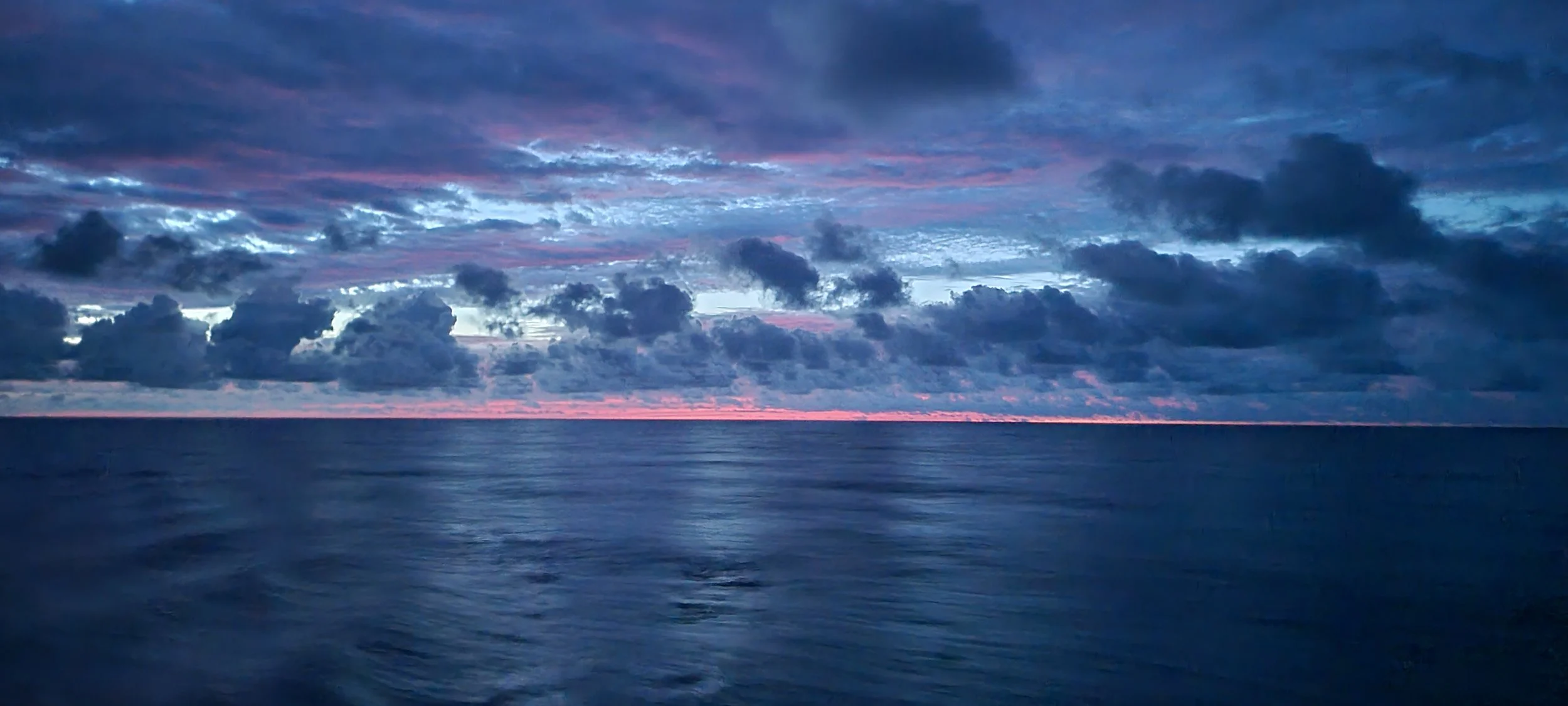 Sky with dark clouds over a calm ocean at sunset or sunrise, with a pink and purple hue near the horizon.