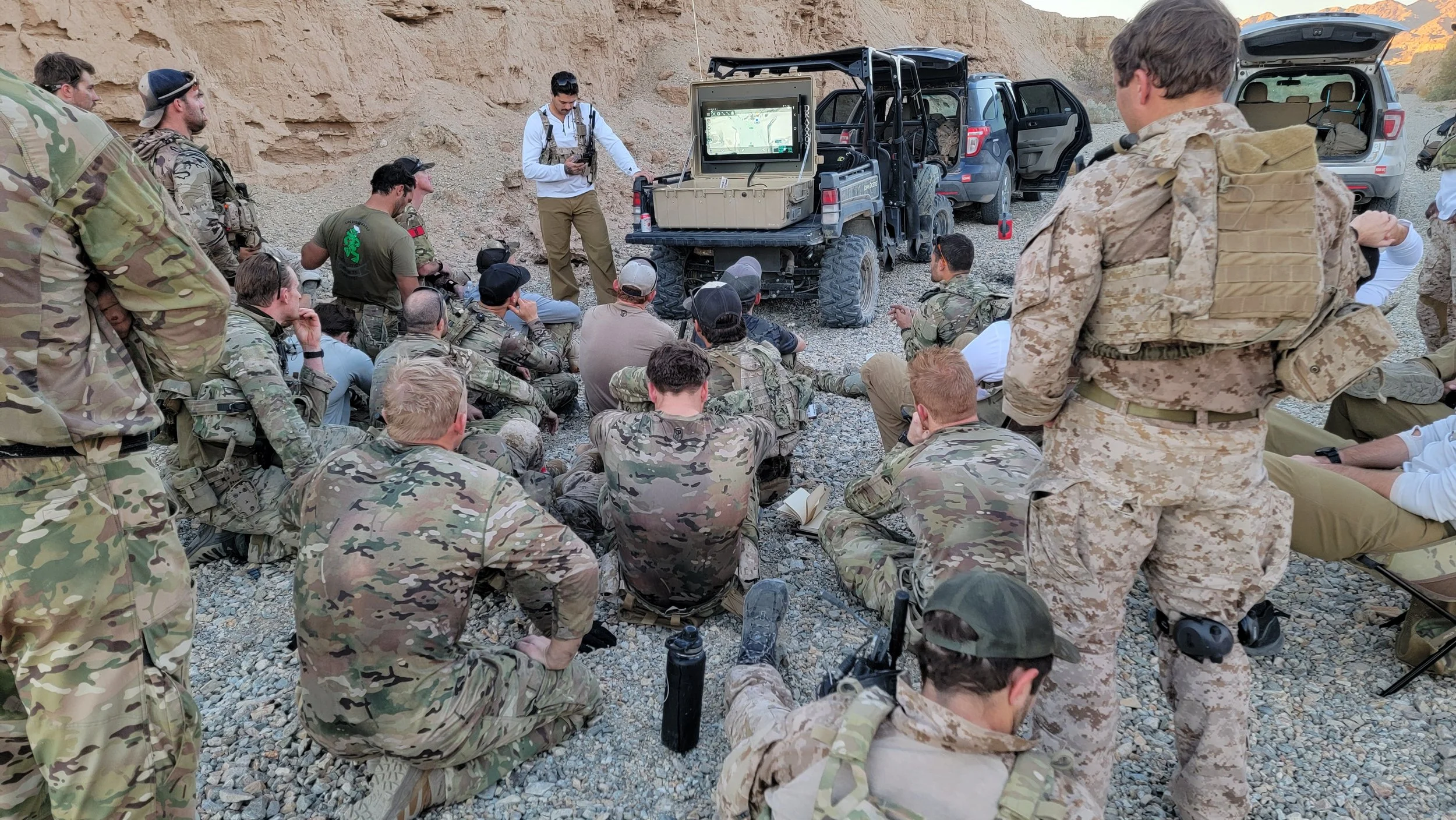 Military personnel in camouflage uniforms gathered outdoors on rocky terrain, listening to a speaker beside military vehicles with open trunks, in a desert landscape.