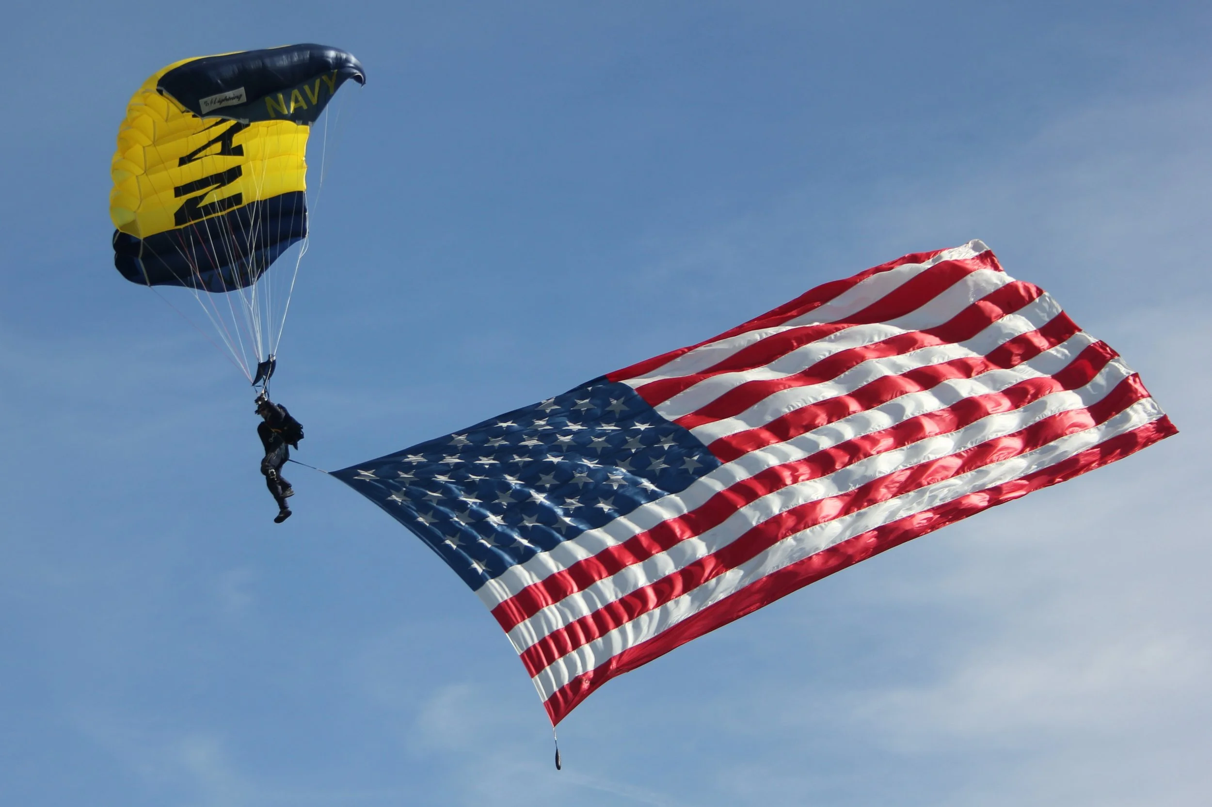 A person skydiving with the American flag and a yellow and black parachute in the sky.
