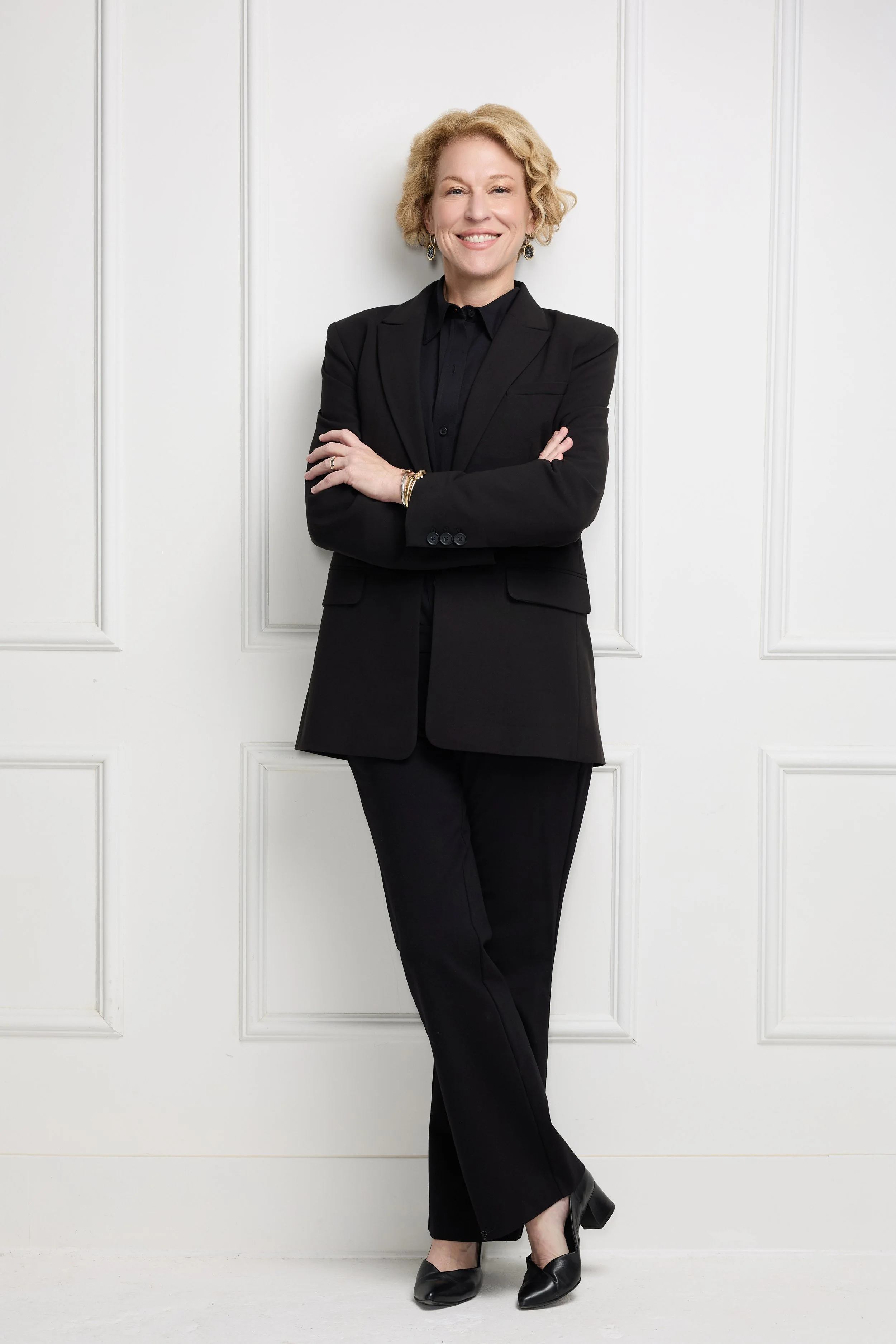 A woman with short curly blonde hair and earrings, smiling with arms crossed, standing against a white paneled wall, wearing a black suit and black heels.