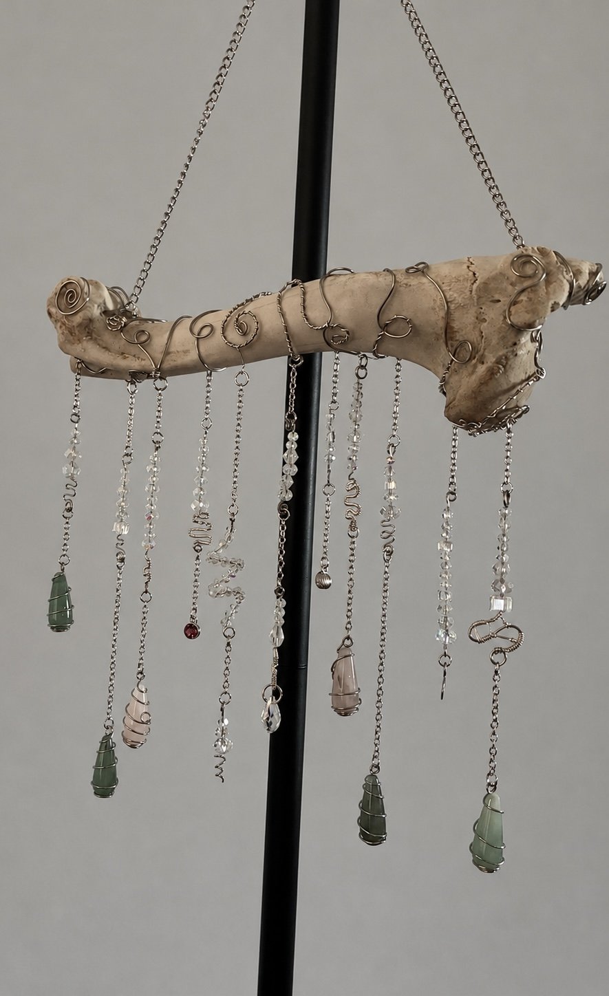 Jewelry display with hanging necklaces and pendants, mounted on a piece of driftwood with metal wire designs, supported by a black stand.
