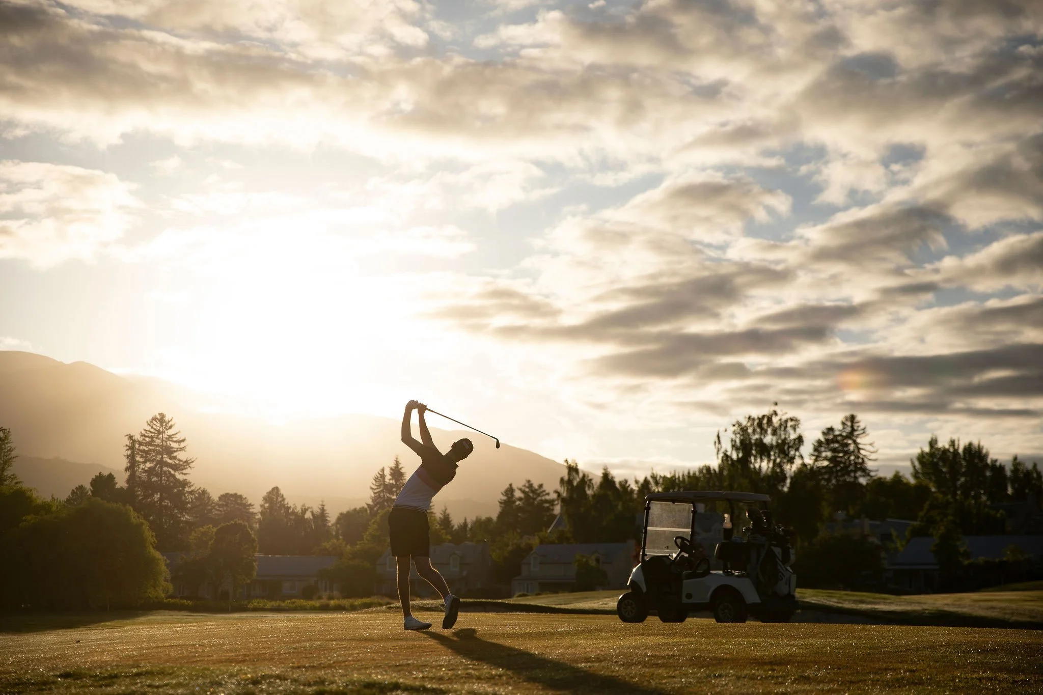 Golfer swinging at sunset on a Queenstown course with a golf cart and mountains in the background.