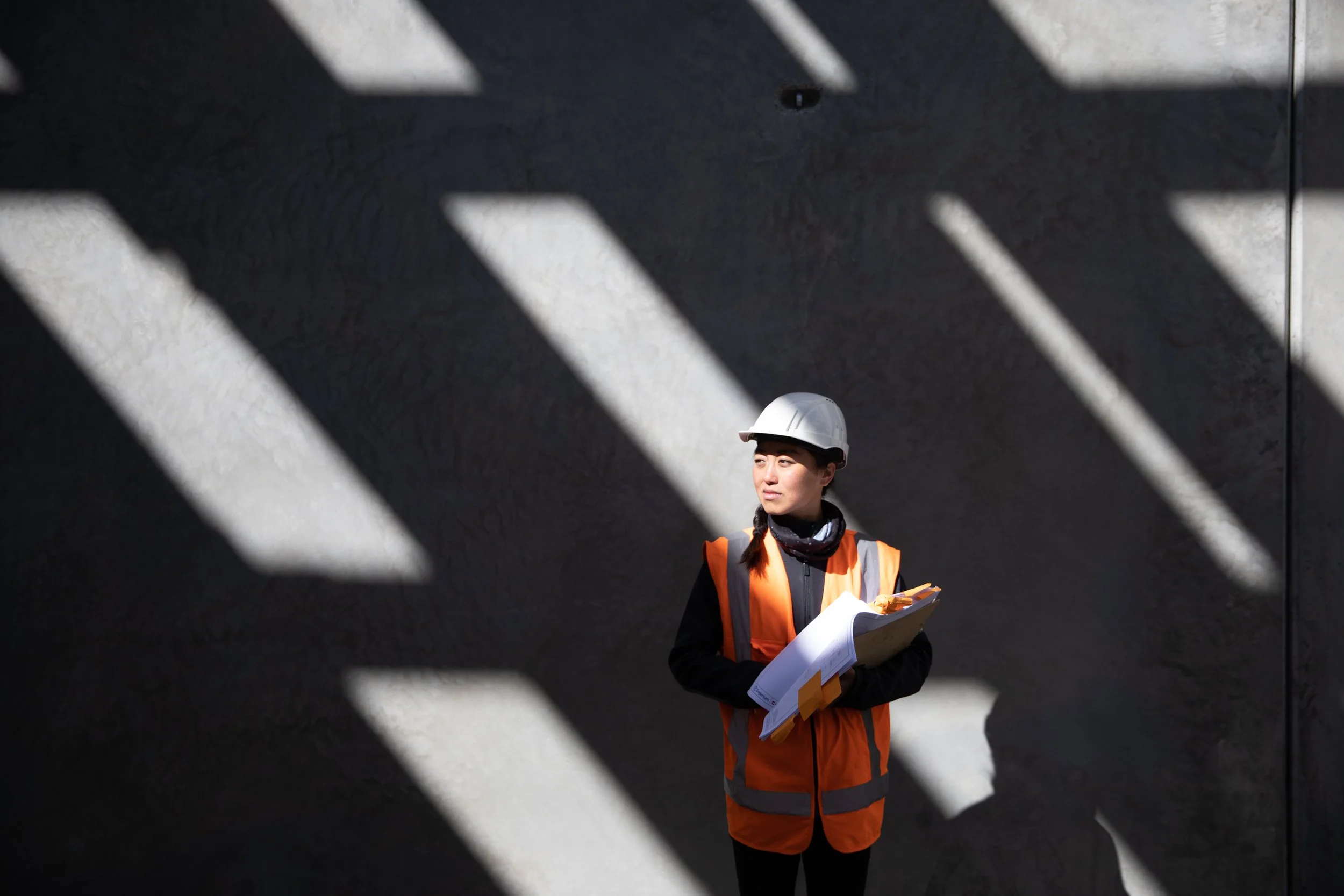 Worker in a hard hat and high-visibility vest photographed in strong architectural shadows.