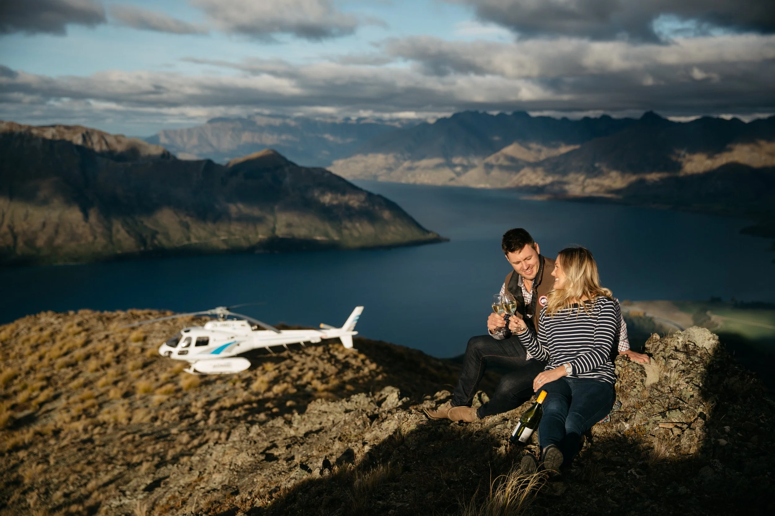 Couple seated beside a helicopter on an alpine ridge above a lake and mountain landscape.