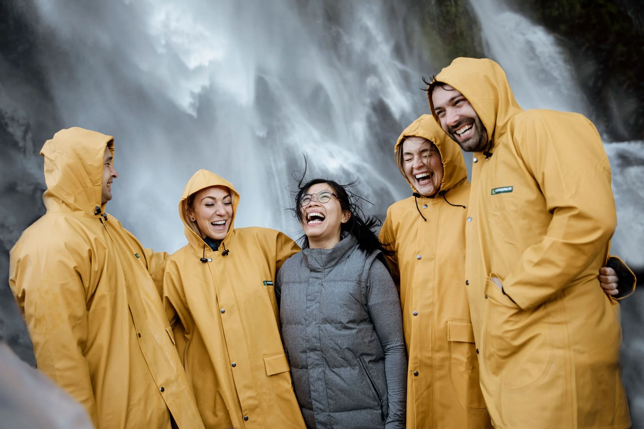 Group of tourists in yellow raincoats laughing beneath a waterfall during a Milford Sound cruise.