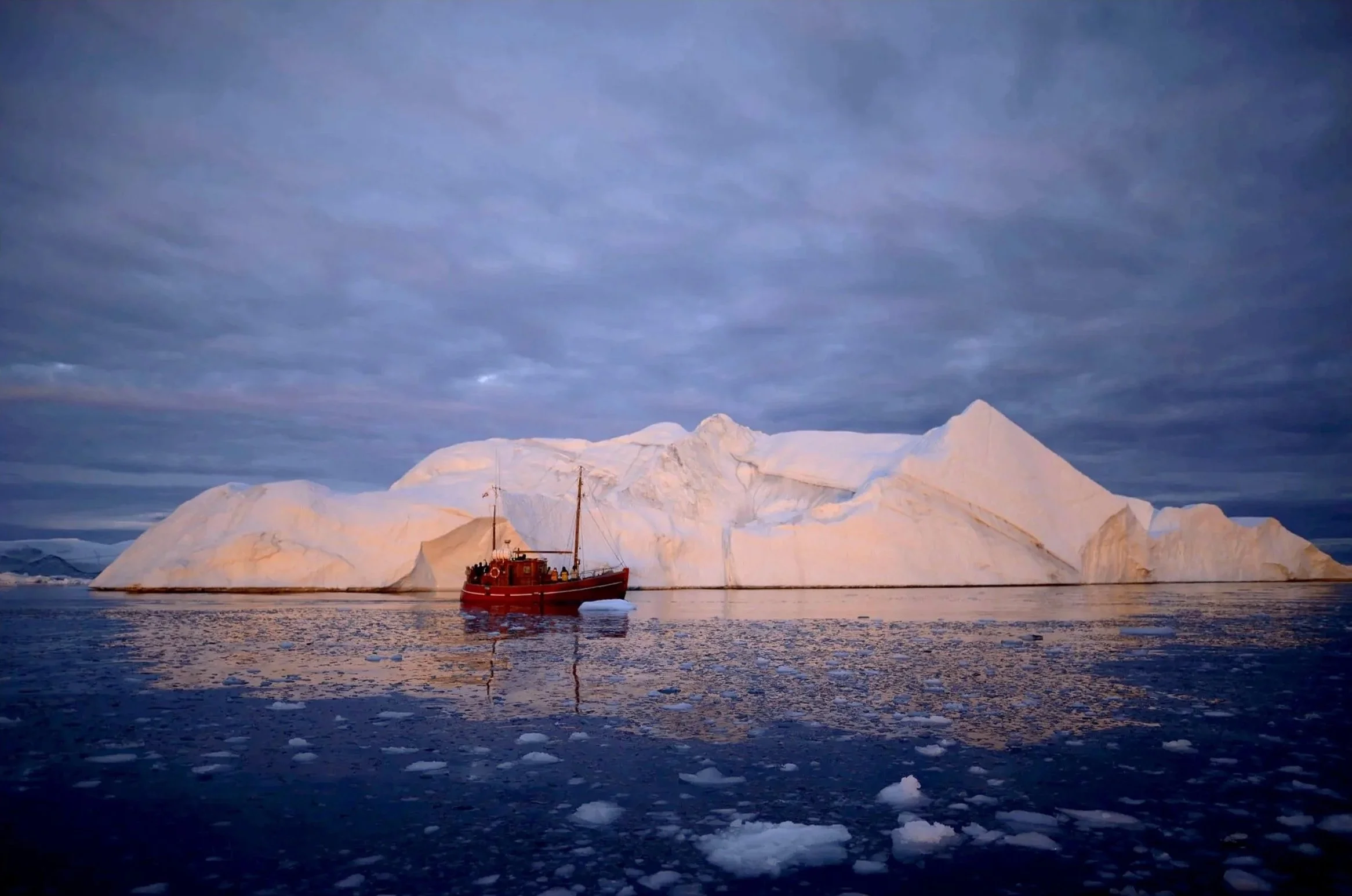Expedition-style landscape photograph showing the scale of a giant iceberg beside a red boat in soft blue evening light.