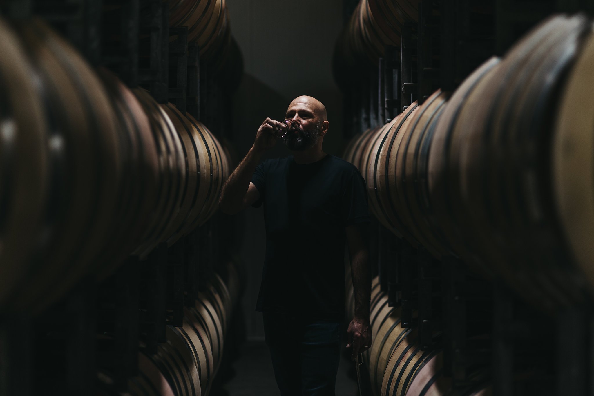 Winemaker standing between rows of barrels in a dark cellar environment.