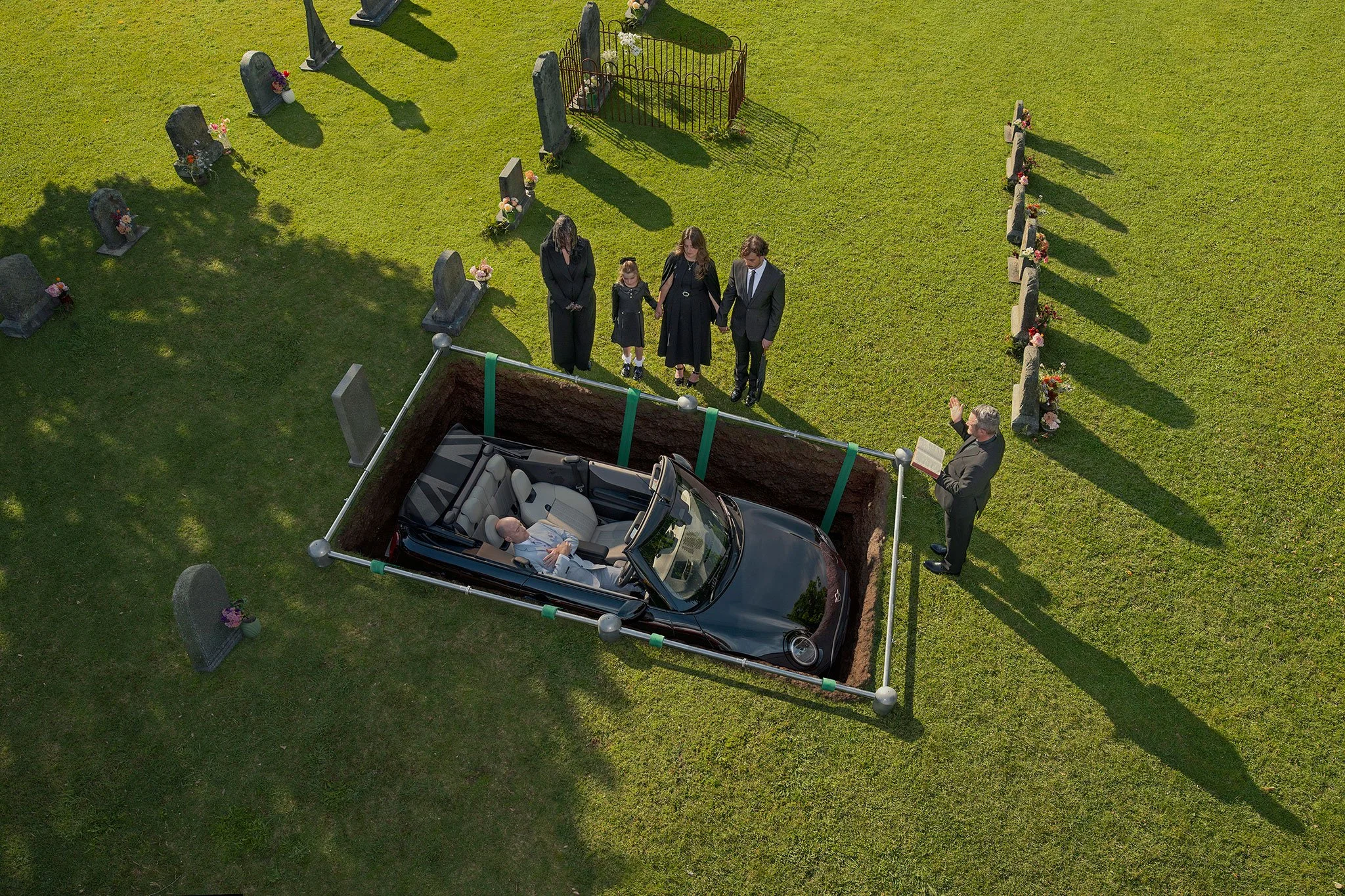 Darkly humorous conceptual image of a funeral scene, with a car lowered into a grave as the central visual idea.