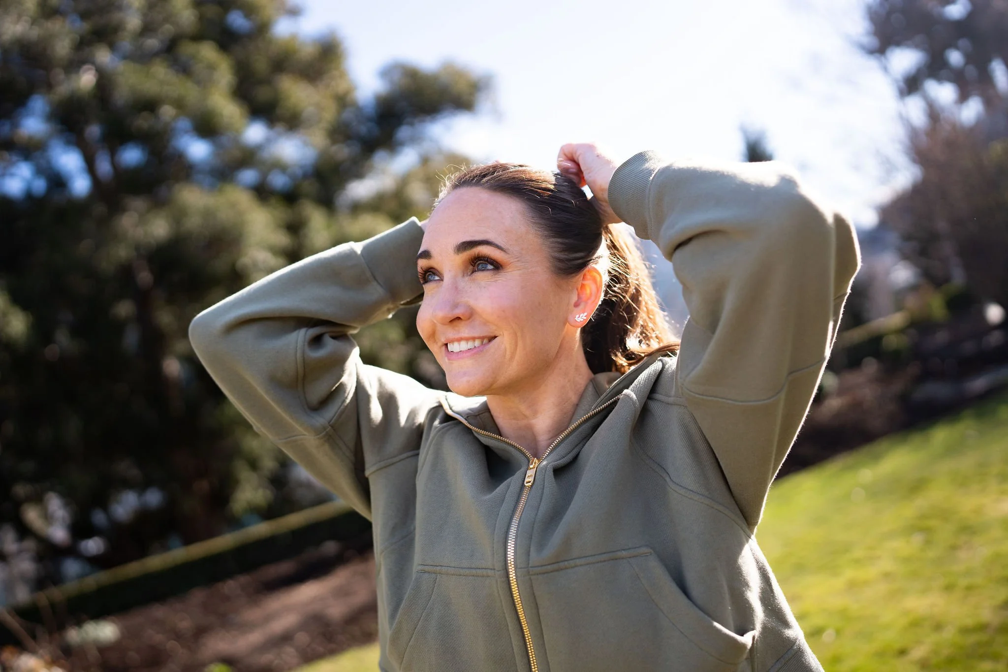 Woman smiling outdoors while tying her hair back in a casual lifestyle portrait.