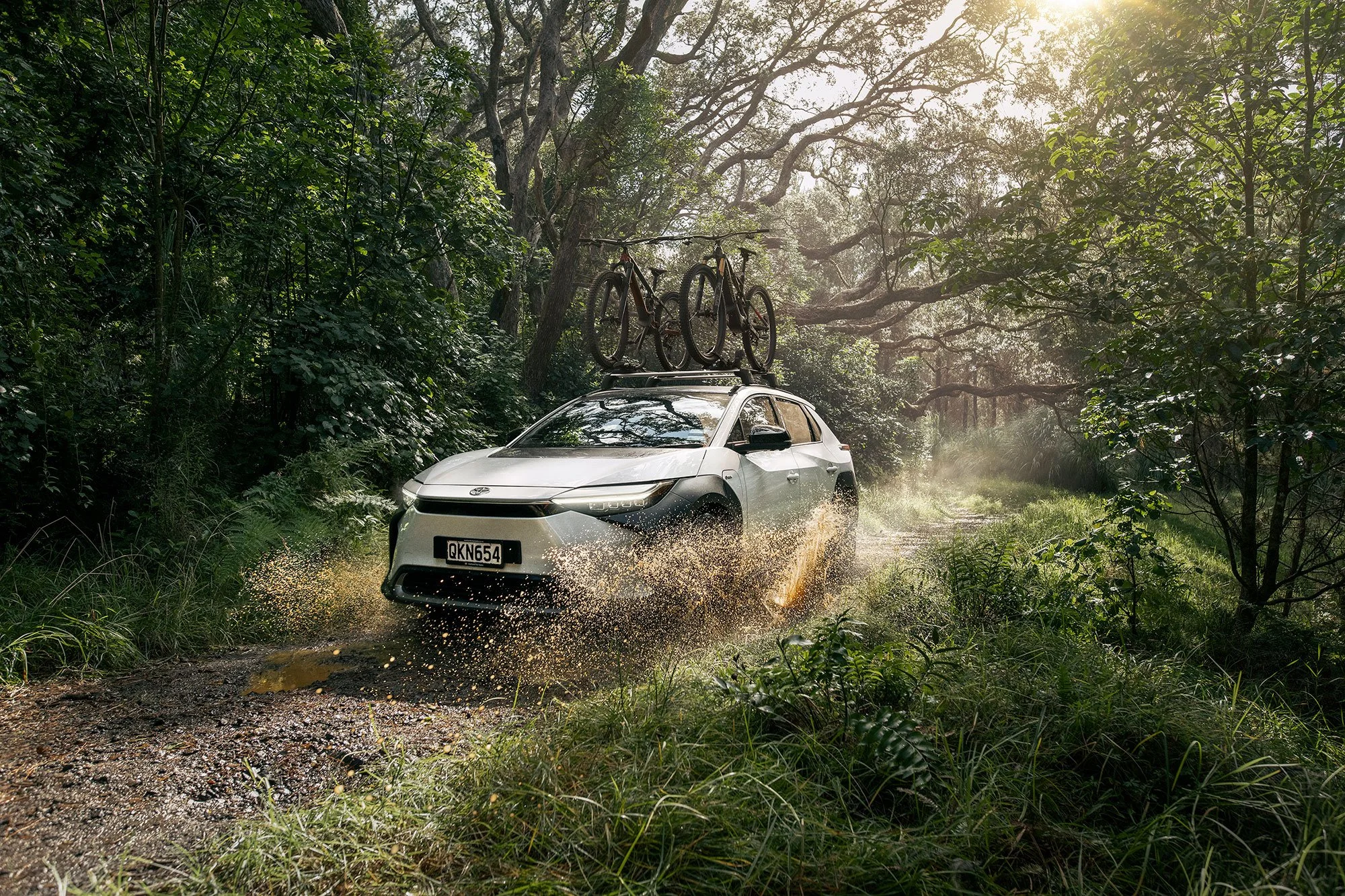 New Zealand automotive campaign image of a Toyota bZ4X moving through a wet forest track, combining electric vehicle photography with cycling and outdoor adventure.