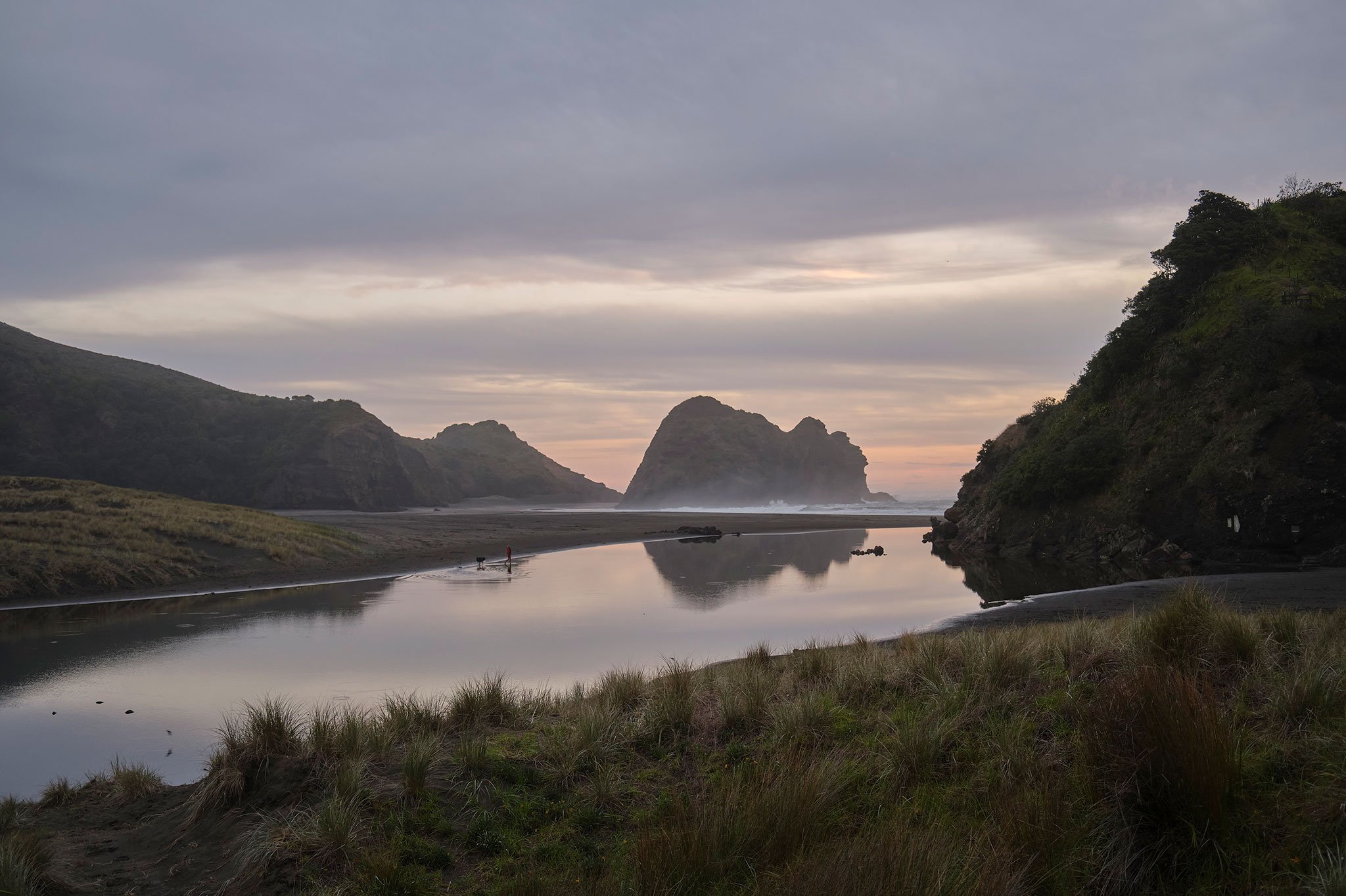 Tranquil coastal landscape photograph of an estuary at low light, with muted sky, reflective water and distant human scale.