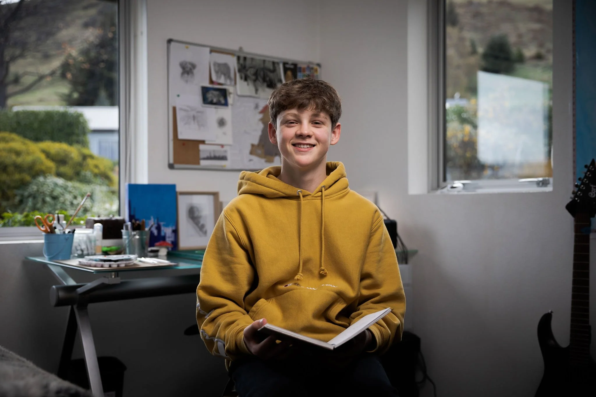 Young artist seated in a studio workspace wearing a yellow hoodie and holding a notebook.