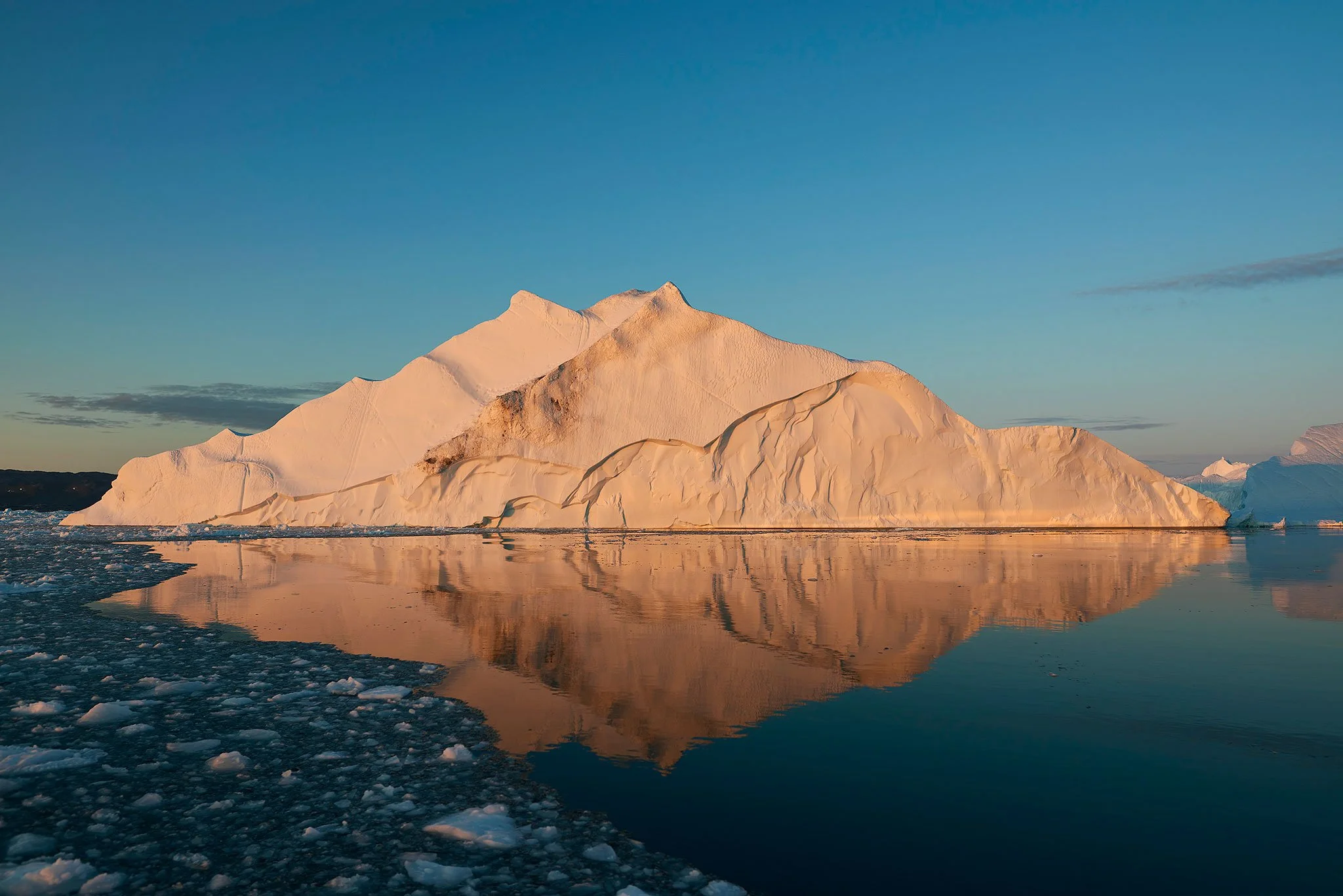Landscape photograph of a sculptural iceberg at golden hour, with still water and a clean reflection creating a quiet, minimalist composition.