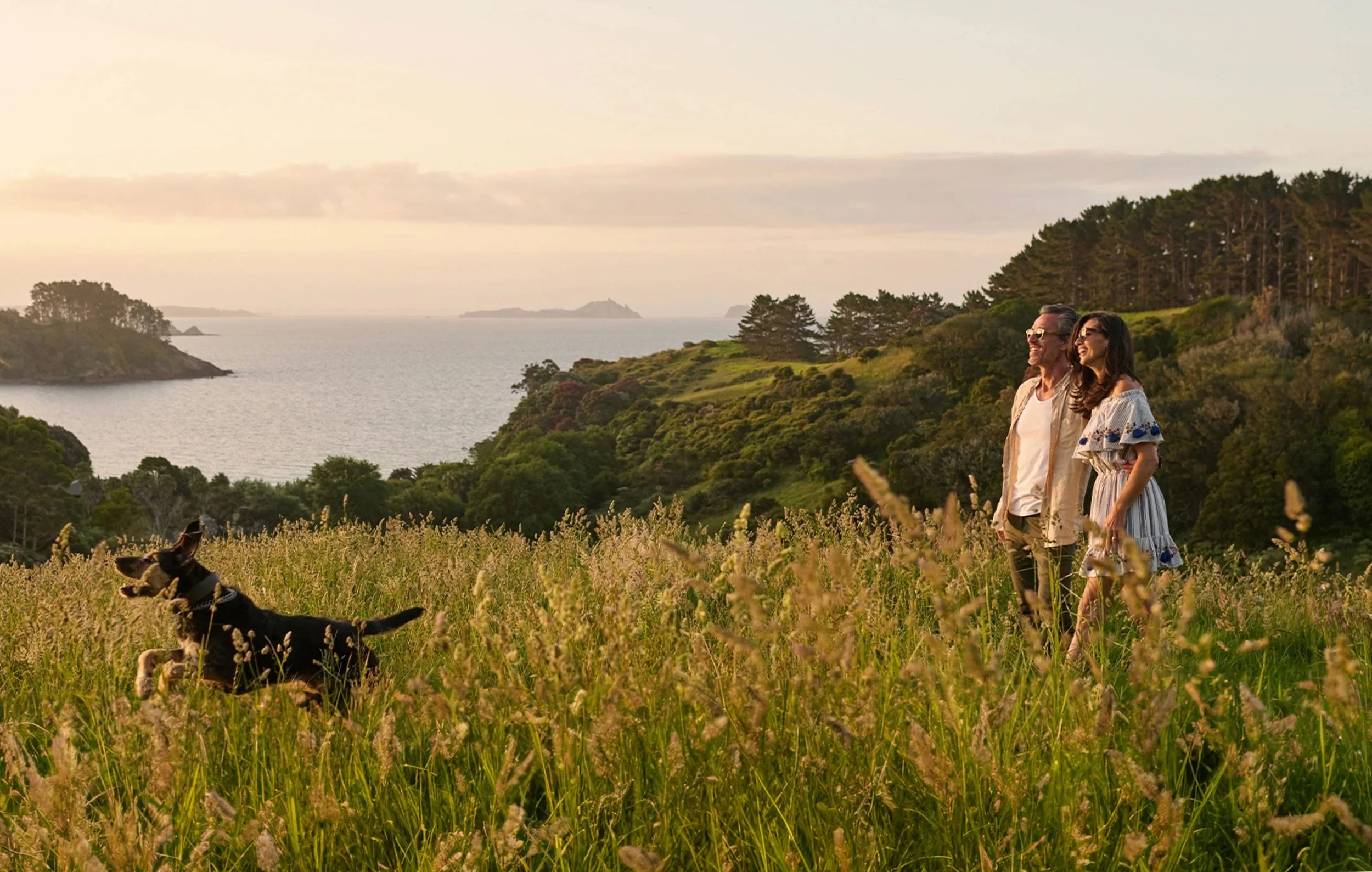 Couple walking with a dog through long grass above a lake at golden hour.