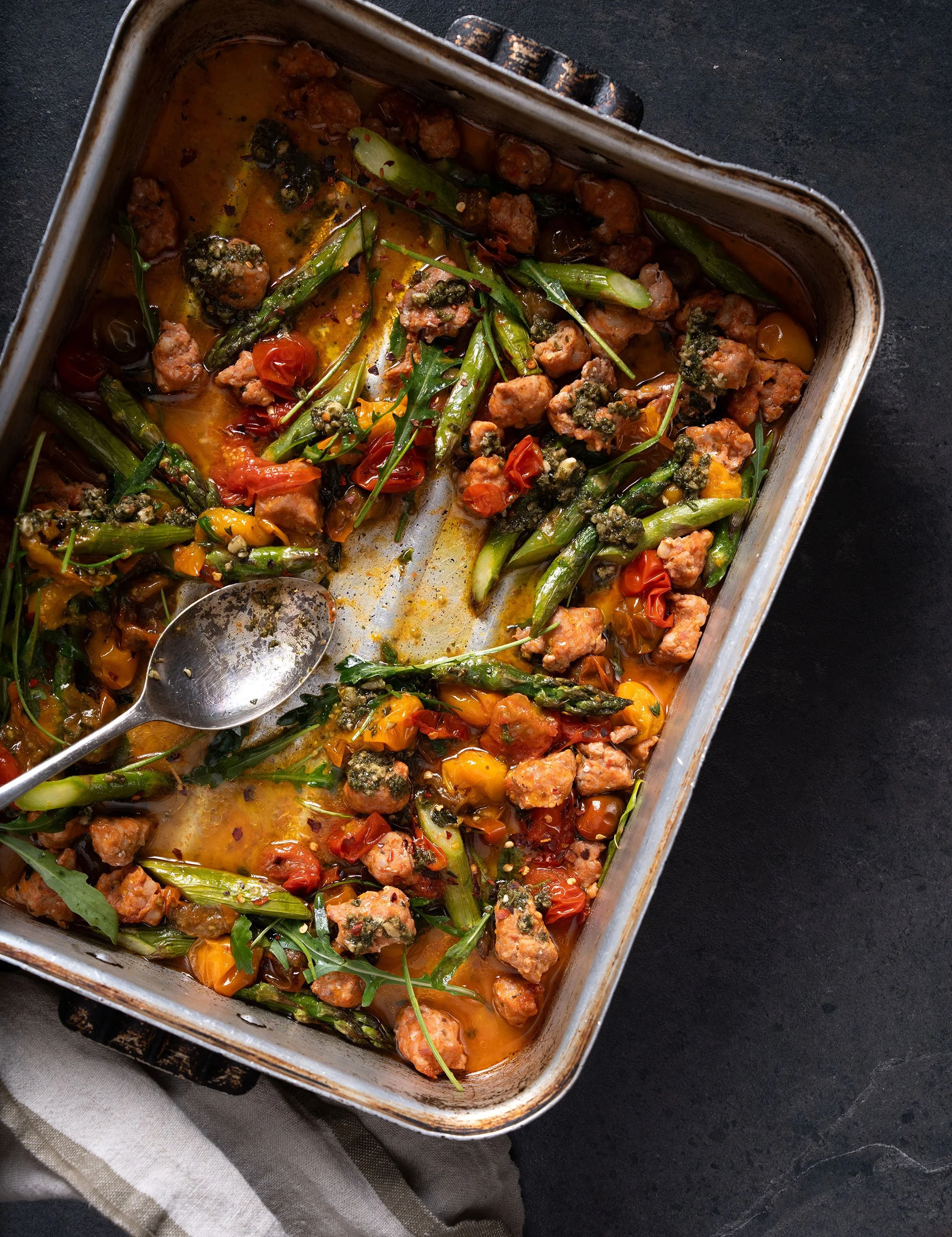 Roasted vegetable tray with herbs and spoon in a dark food photography setup.