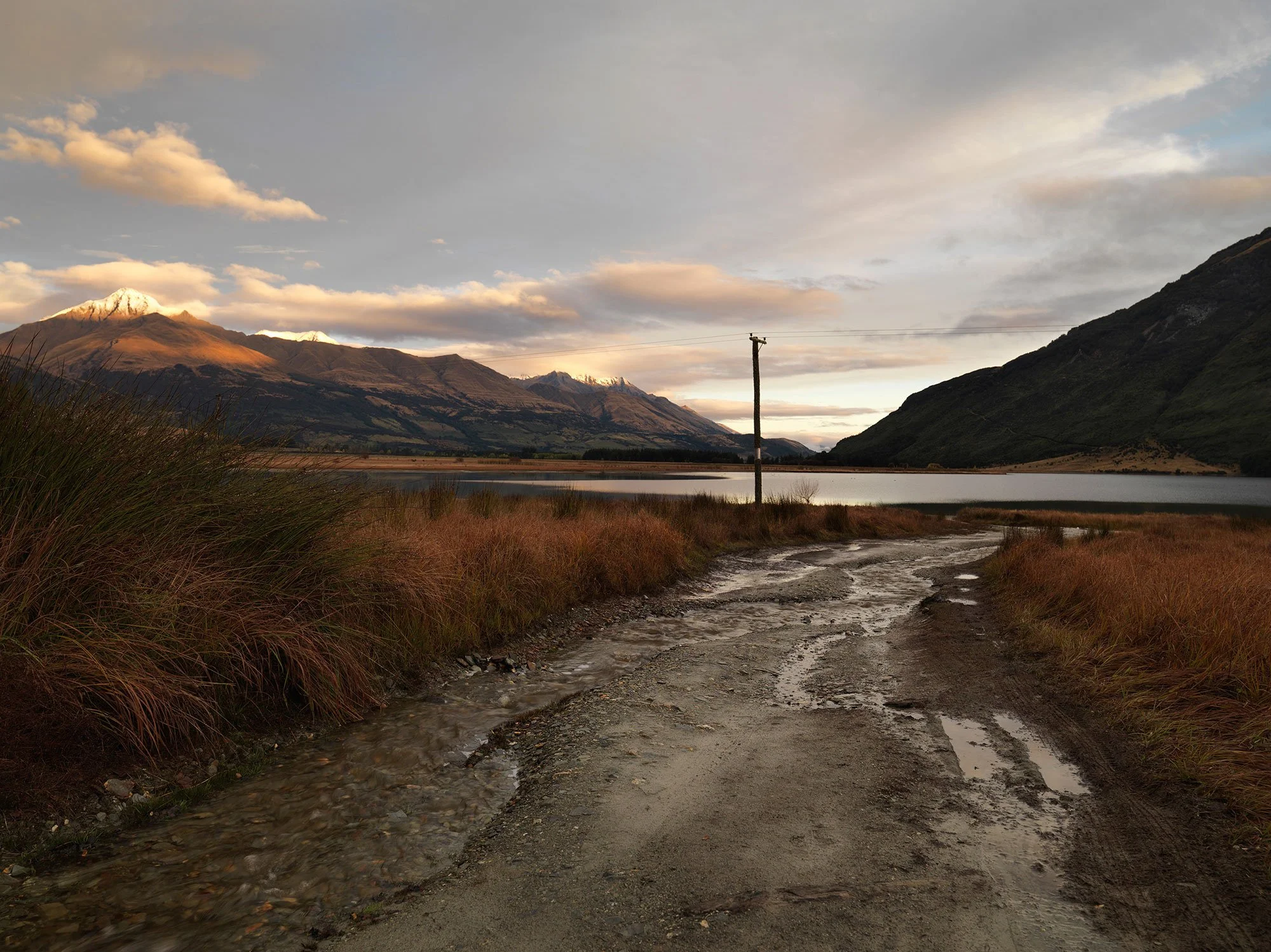 Landscape photograph of a rural alpine scene with textured foreground, still lake and warm light touching distant peaks.