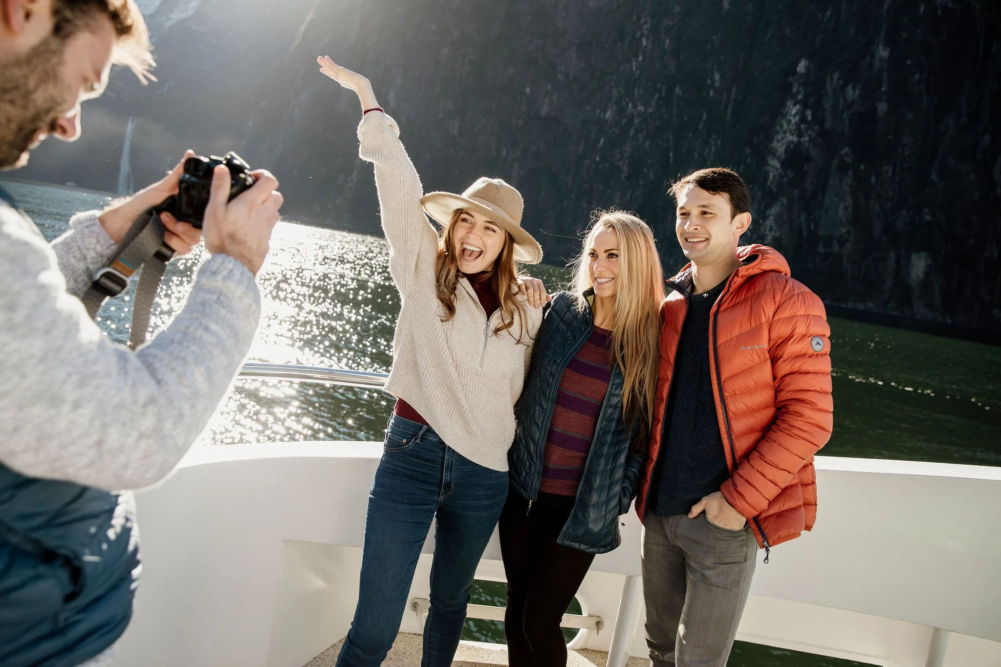 Tourists being photographed on a boat deck during a Milford Sound cruise beneath steep cliffs.