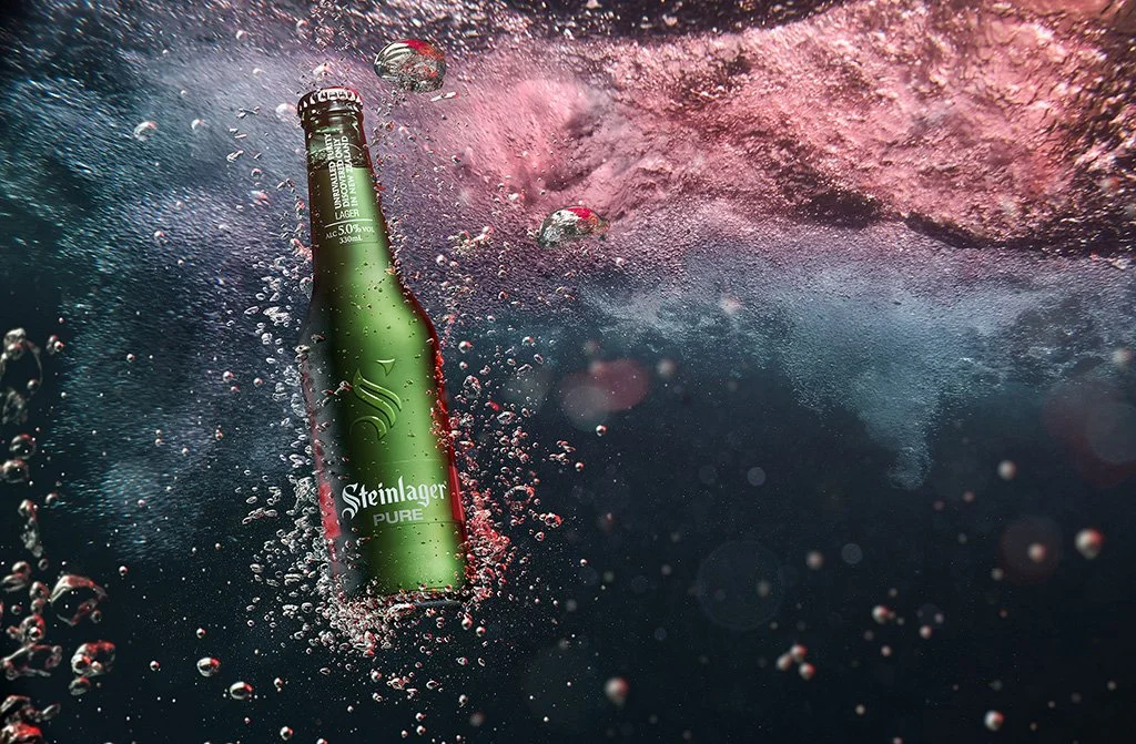 Steinlager bottle suspended in a vivid storm of pink and blue water for a conceptual product image.