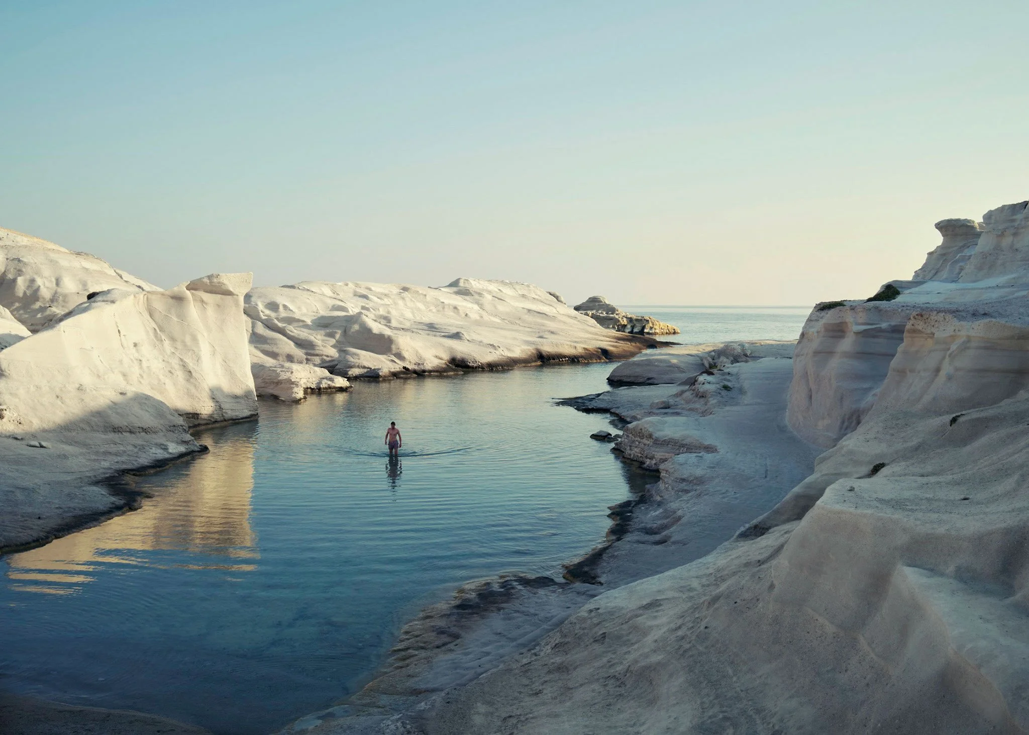 Coastal landscape photograph of a lone swimmer in a serene natural pool framed by pale sculpted rock and still water.